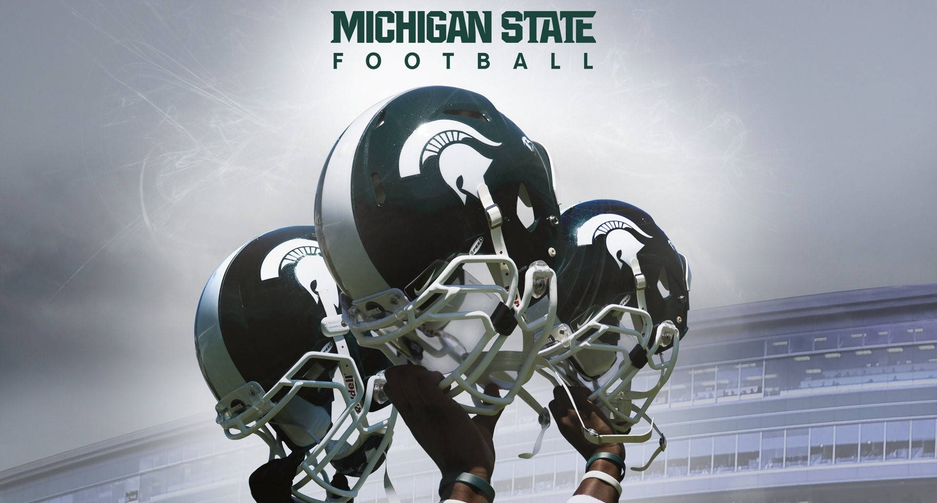 Michigan State Spartans 桌布 壁紙 — 三個密西根州立大學美式足球頭盔在暴風雨天空下，於戲劇性的體育場背景下高舉。