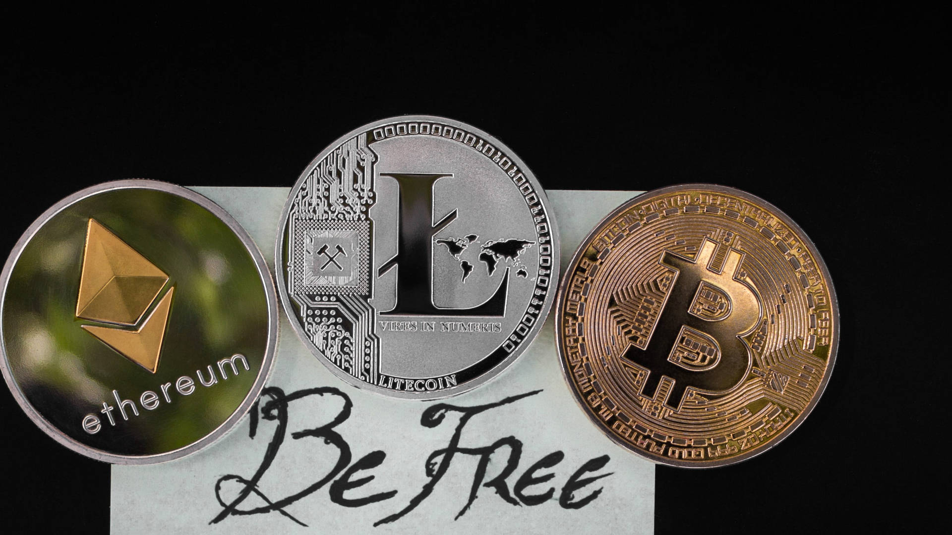 以太坊 桌布 壁紙 — 三個實體加密貨幣硬幣比特幣、以太坊和萊特幣放在一張紙上，紙上寫有手寫的「Be Free」，背景為黑色。