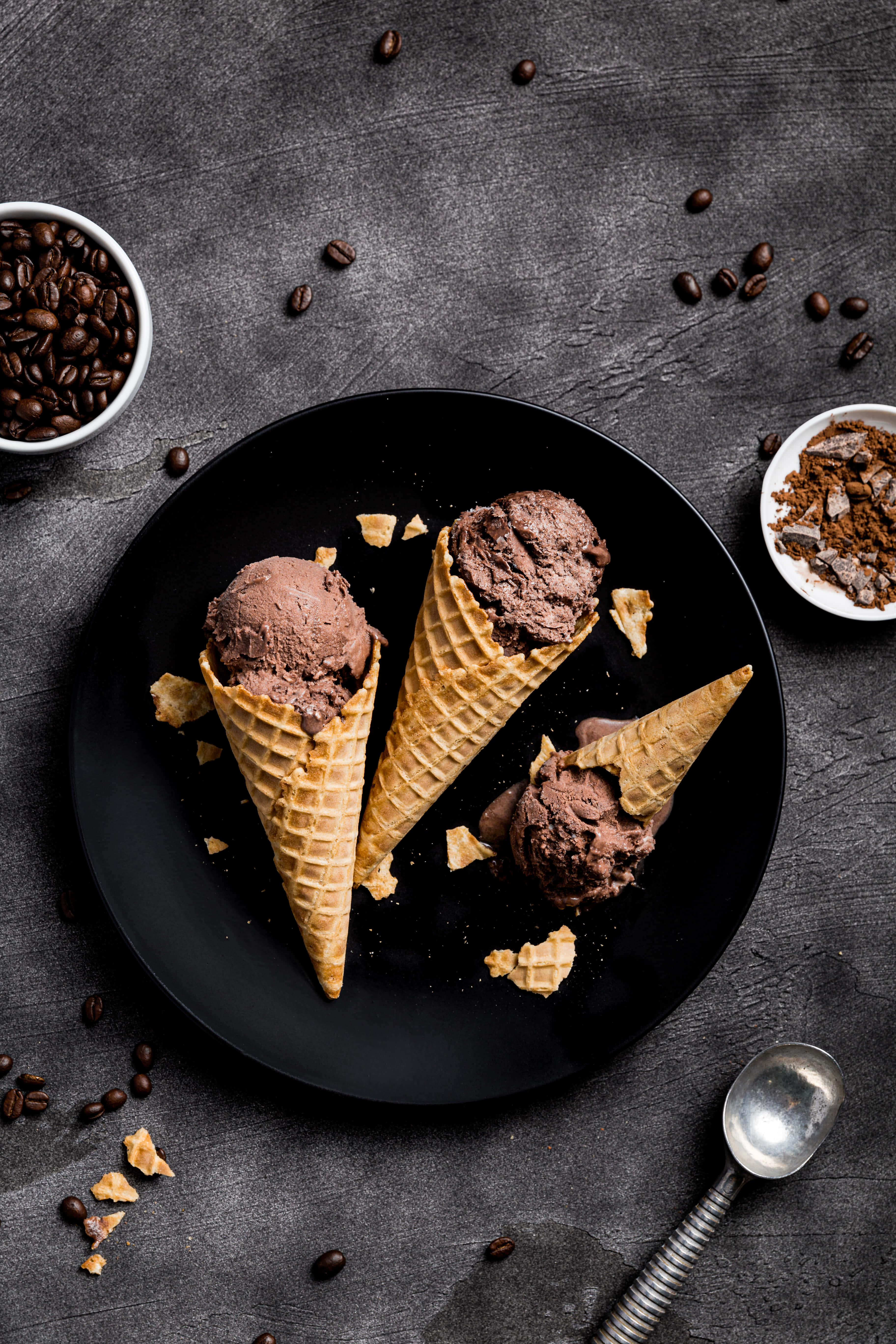 Ice Cream 桌布 壁紙 — 三個巧克力冰淇淋甜筒放在黑色盤子上，周圍有咖啡豆和碎鬆餅塊，置於深色紋理表面。