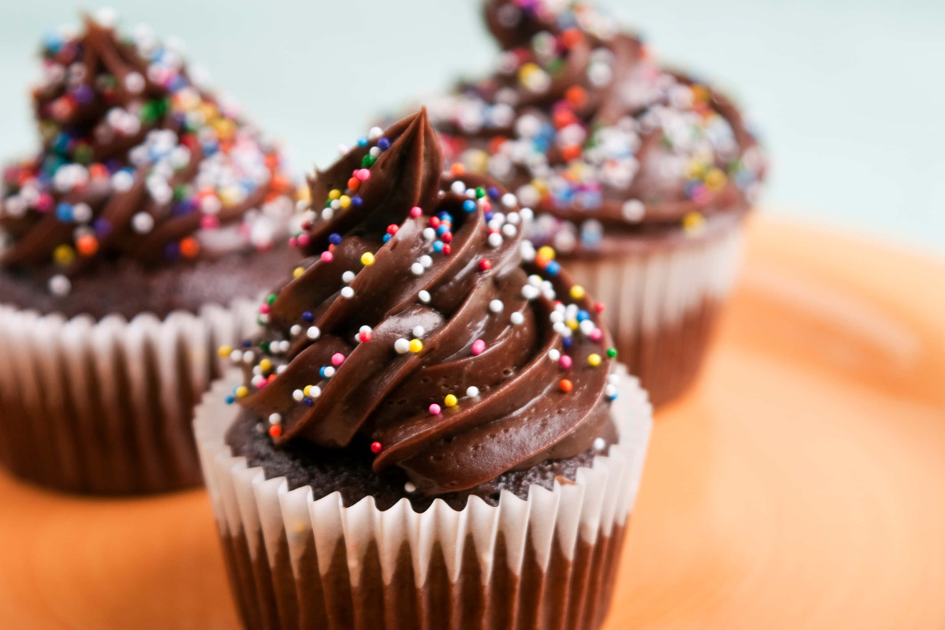 Cute Cupcake 桌布 壁紙 — 三個巧克力紙杯蛋糕，上面有濃郁的糖霜漩渦和色彩繽紛的糖粒，放在橙色盤子上。