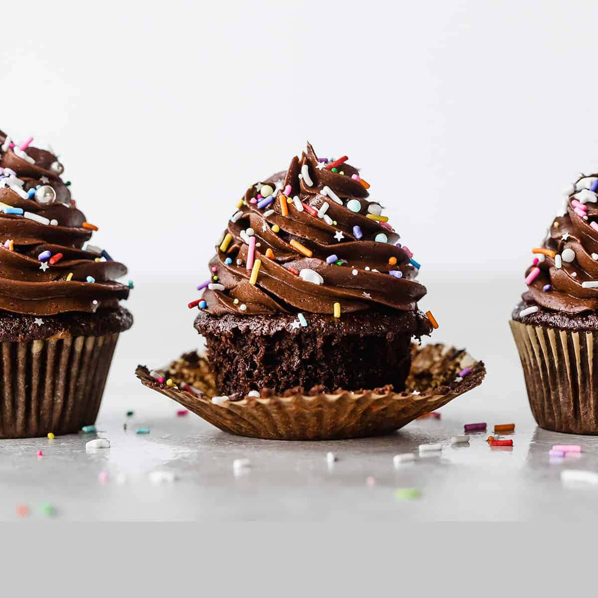 Cupcake 桌布 壁紙 — 三個巧克力紙杯蛋糕，帶有漩渦糖霜和彩色糖粒，放在灰色表面上，周圍散落著糖粒。
