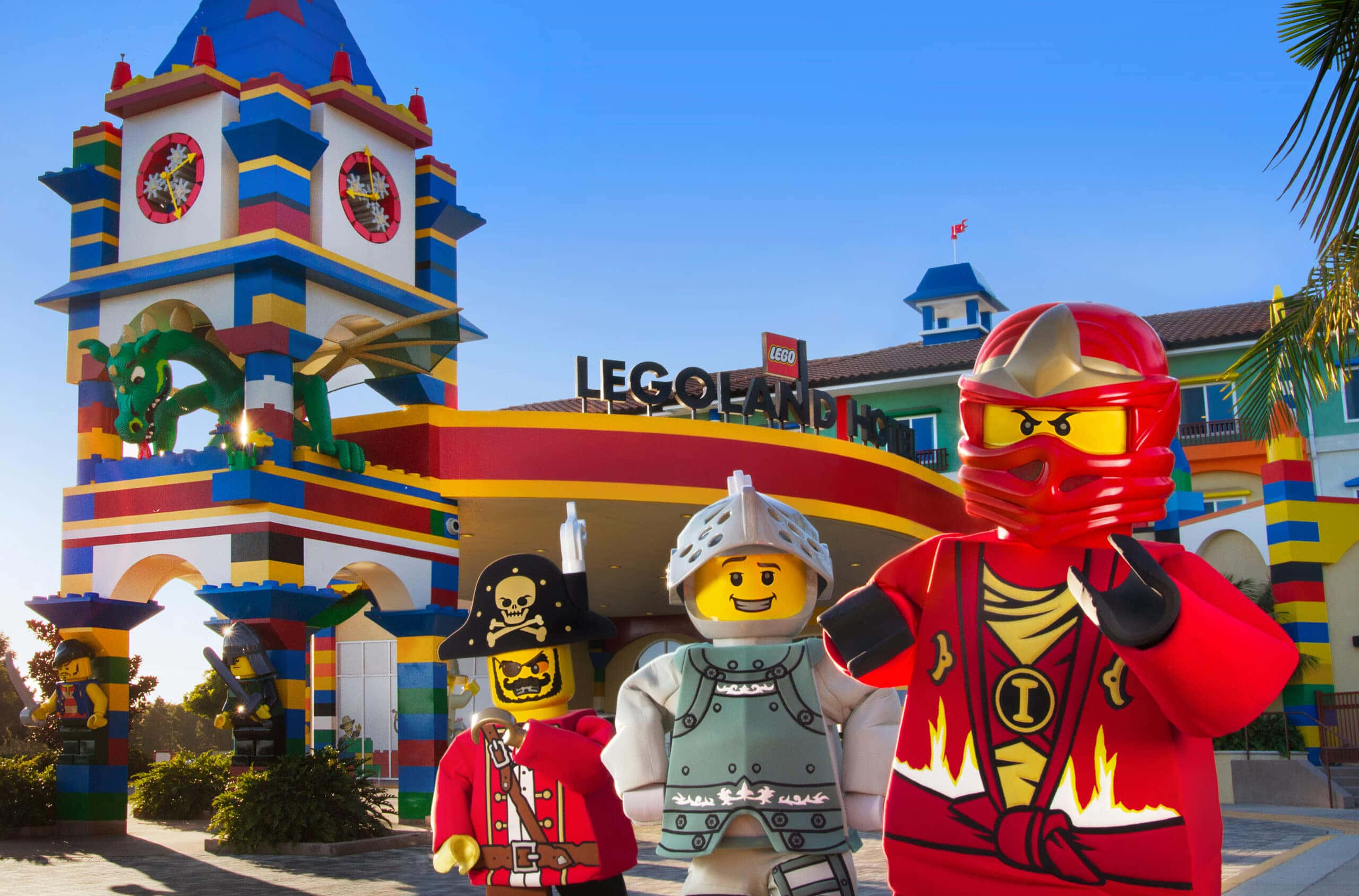 Legoland 桌布 壁紙 — 三個巨型 LEGO 人偶自豪地站在色彩繽紛的 LEGO Land Hotel 前，背景是晴朗的藍天。
