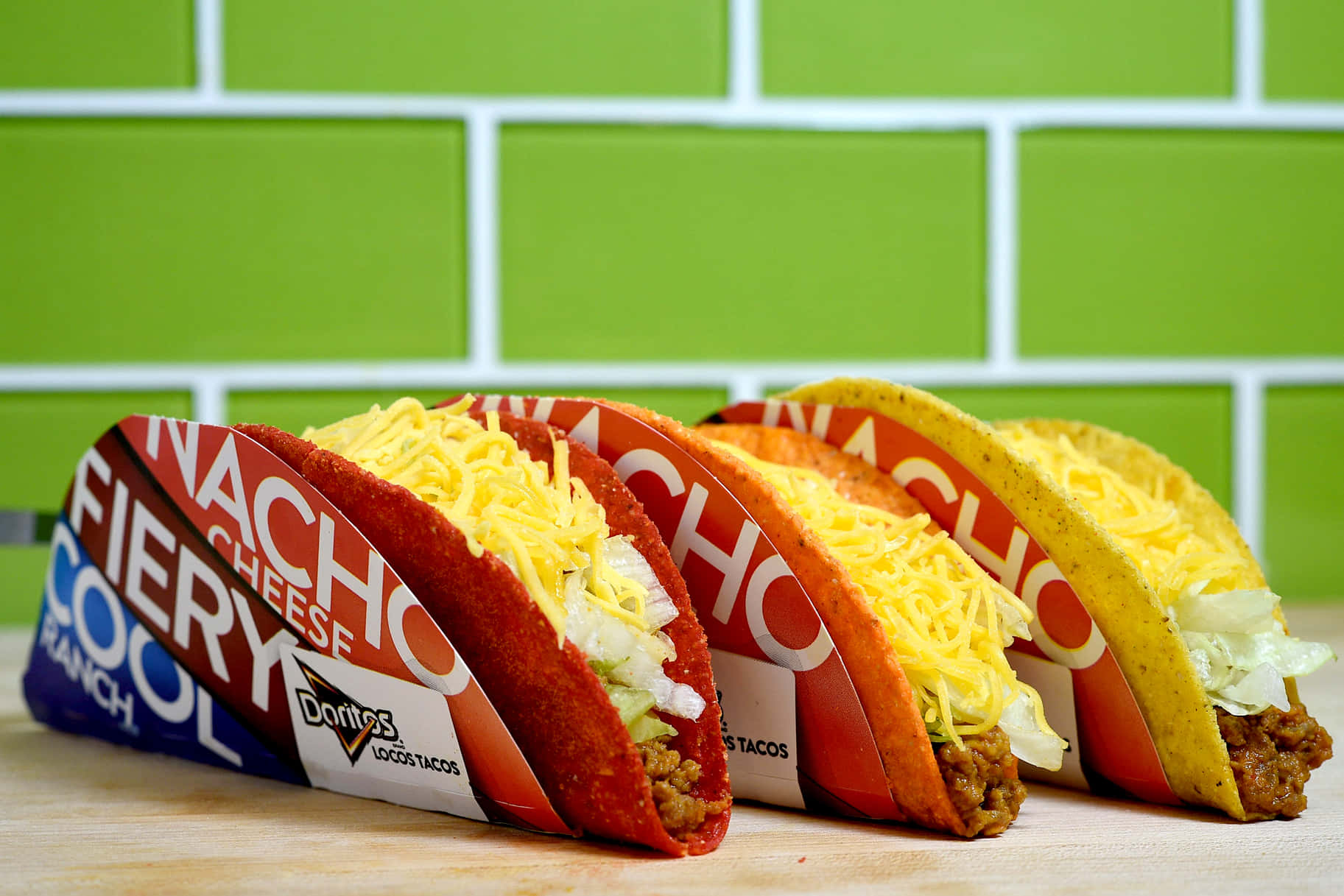 塔可鐘 桌布 壁紙 — 三個帶有品牌外殼的Doritos Locos Tacos放在木質表面上，背景是明亮的綠色瓷磚牆。