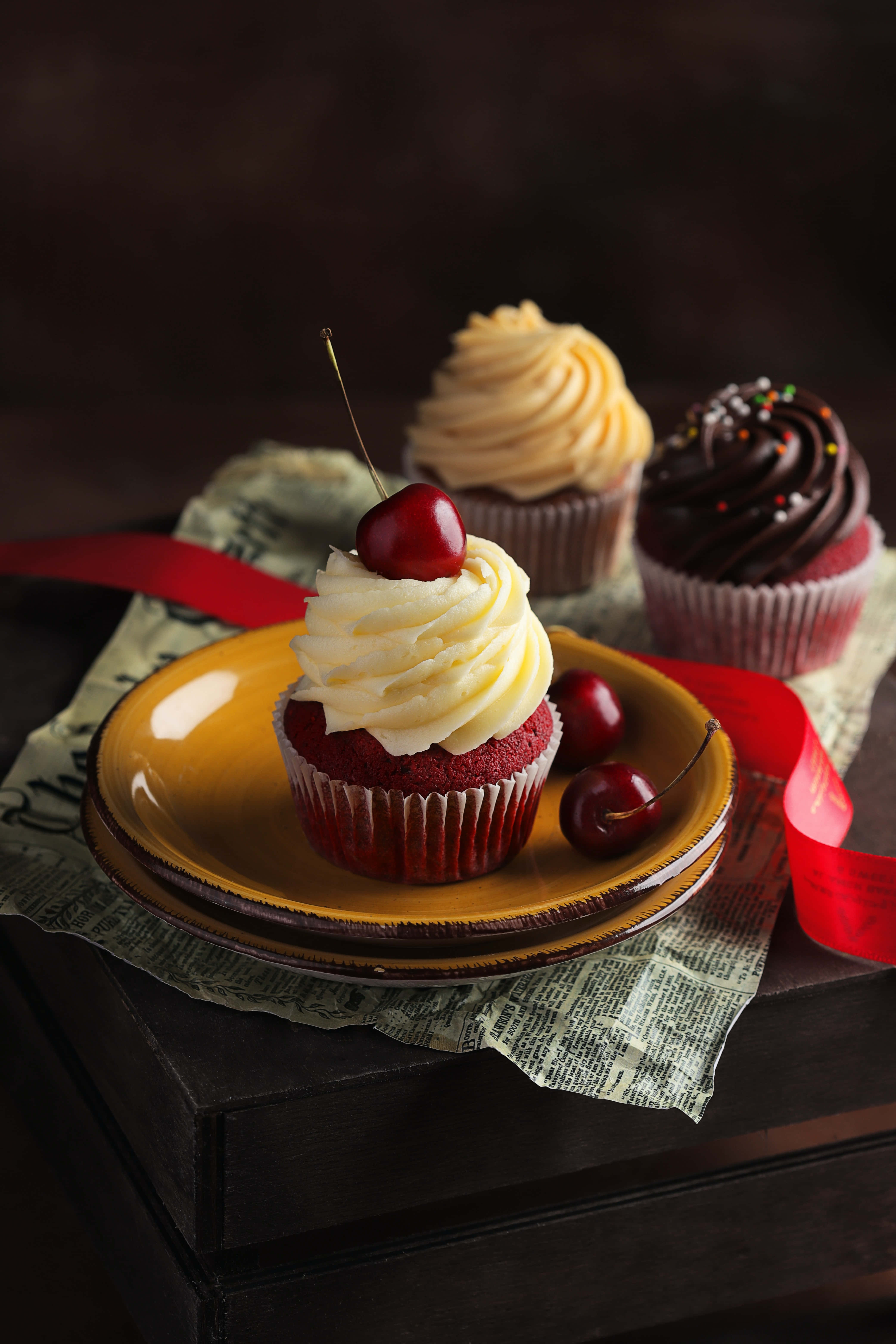 Cupcake 桌布 壁紙 — 三個帶有奶油乳酪糖霜和櫻桃的頹廢紙杯蛋糕，放在復古黃色盤子上，盤子下墊著報紙和紅色絲帶。