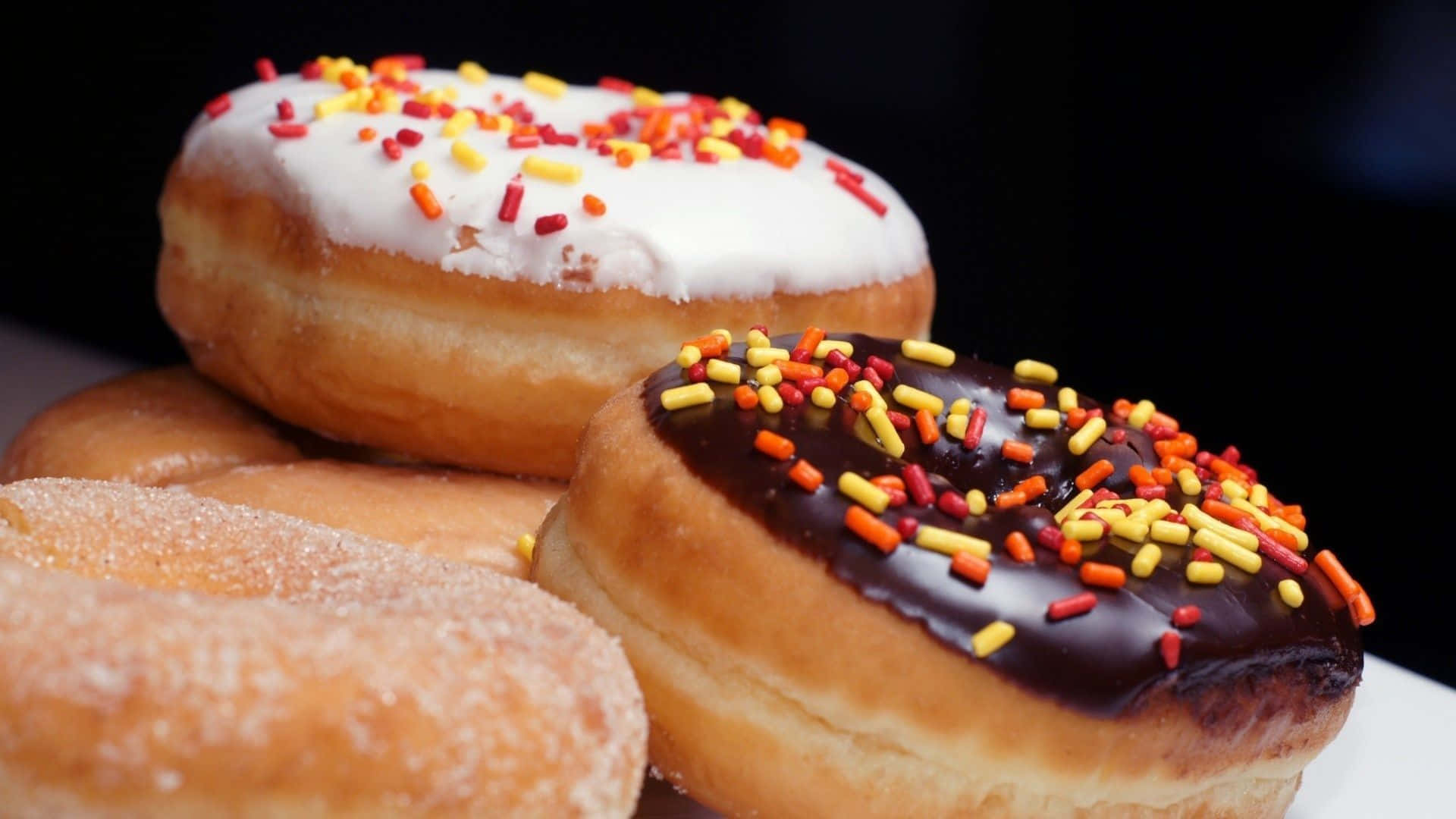 donuts 桌布 壁紙 — 三個帶有彩色糖粒和糖衣的釉面甜甜圈堆疊在白色盤子上，背景為深色。