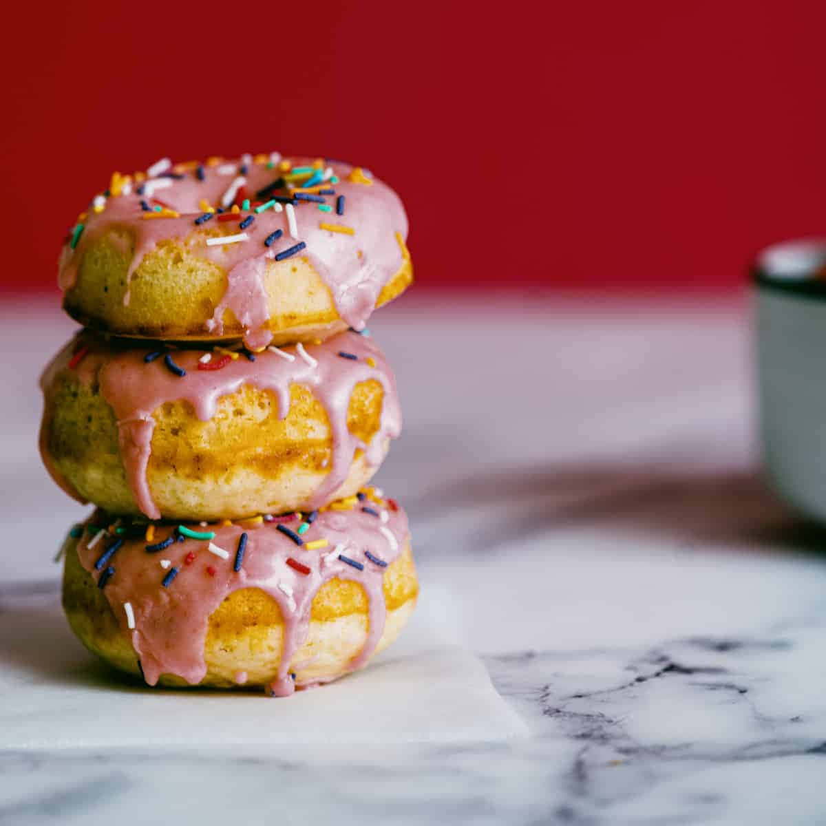 Glazed Donut 桌布 壁紙 — 三個帶有彩色糖粒的釉面甜甜圈堆疊在大理石表面上，背景為大膽的紅色。