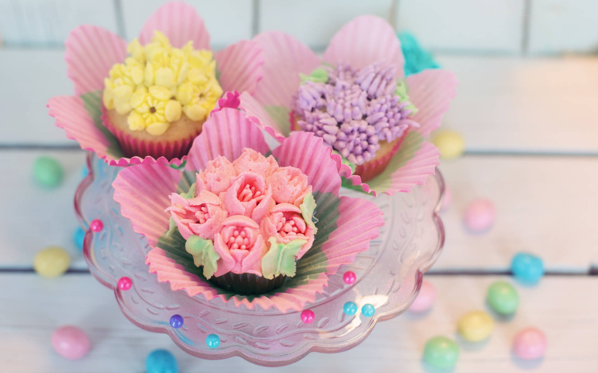 pastel 桌布 壁紙 — 三個帶有粉紅色、黃色和紫色花朵形狀糖霜的杯子蛋糕放在玻璃架上，周圍環繞著色彩繽紛的糖果。