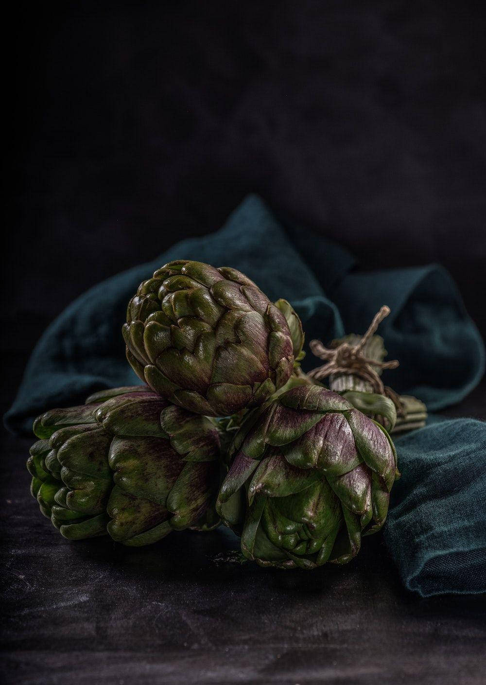 Food Photography 桌布 壁紙 — 三個帶有綠色和紫色色調的朝鮮薊放在深青綠色的布上，背景為黑色。