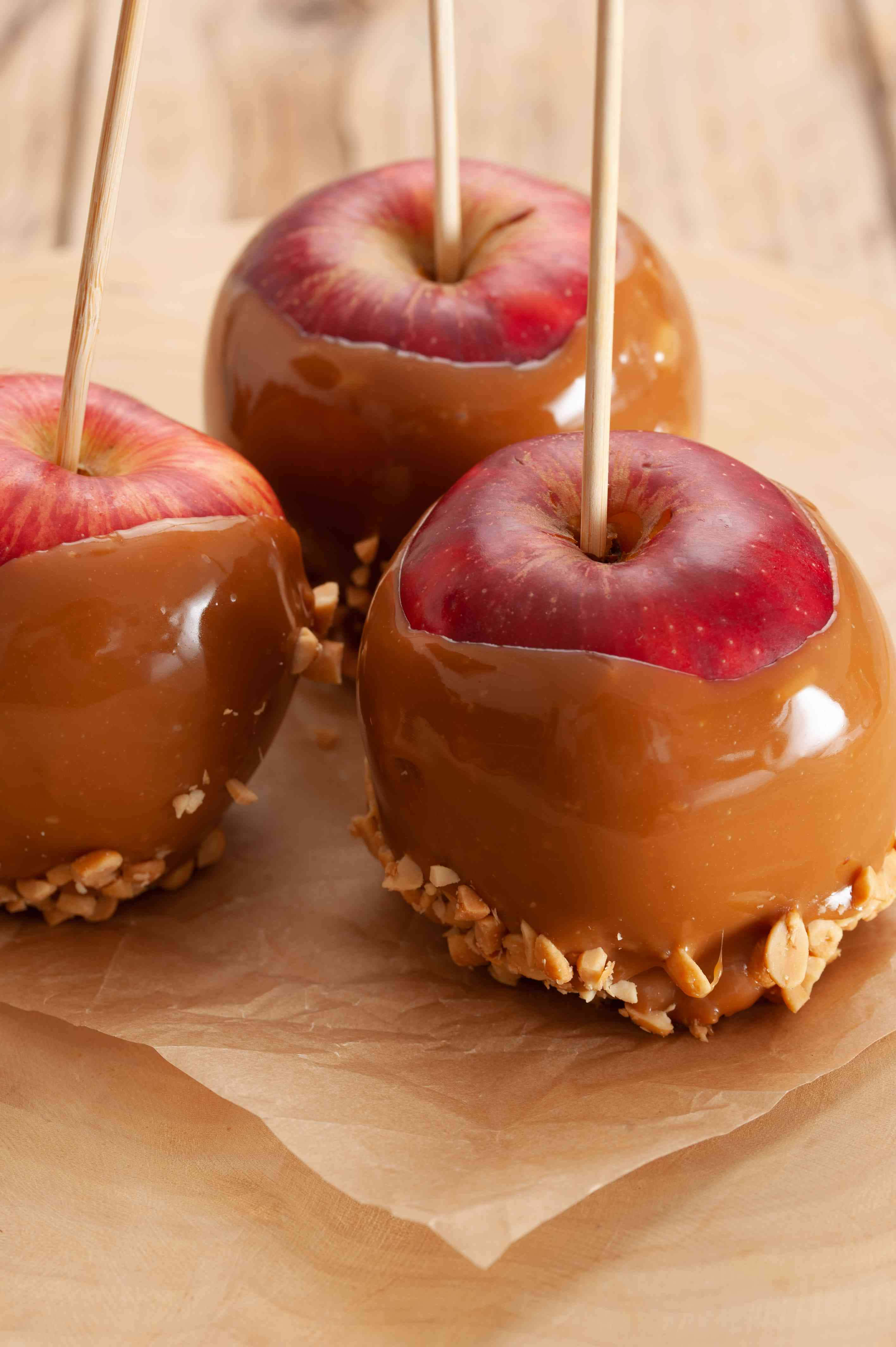 Caramel Apples 桌布 壁紙 — 三個帶有花生配料的光澤焦糖蘋果放在木質表面上的棕色烘焙紙上。
