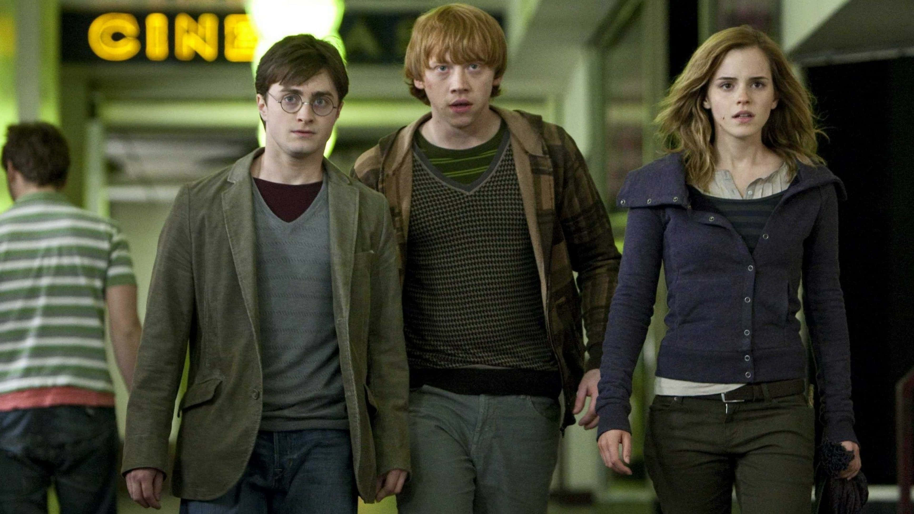Harry Potter 4K 桌布 壁紙 — 三個年輕人走在一條燈光昏暗的走廊上，上方有一個發光的電影院標誌。