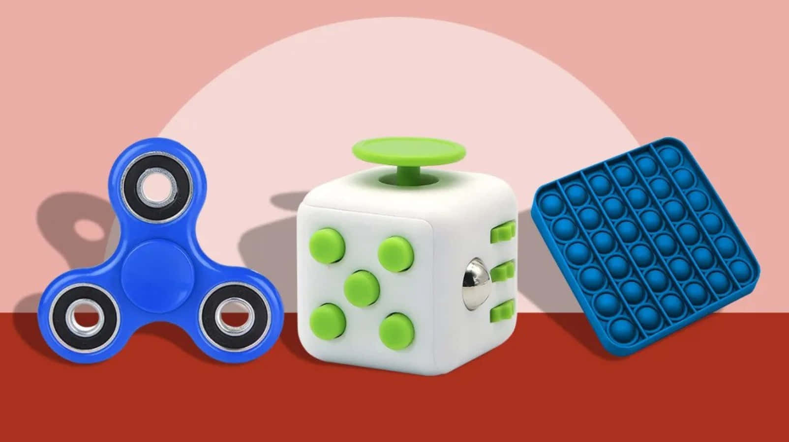 Fidget 桌布 壁紙 — 三個彩色減壓玩具：一個藍色旋轉器、一個帶綠色按鈕的白色立方體和一個藍色泡泡墊，放在紅色表面上，背景為柔和粉紅色。
