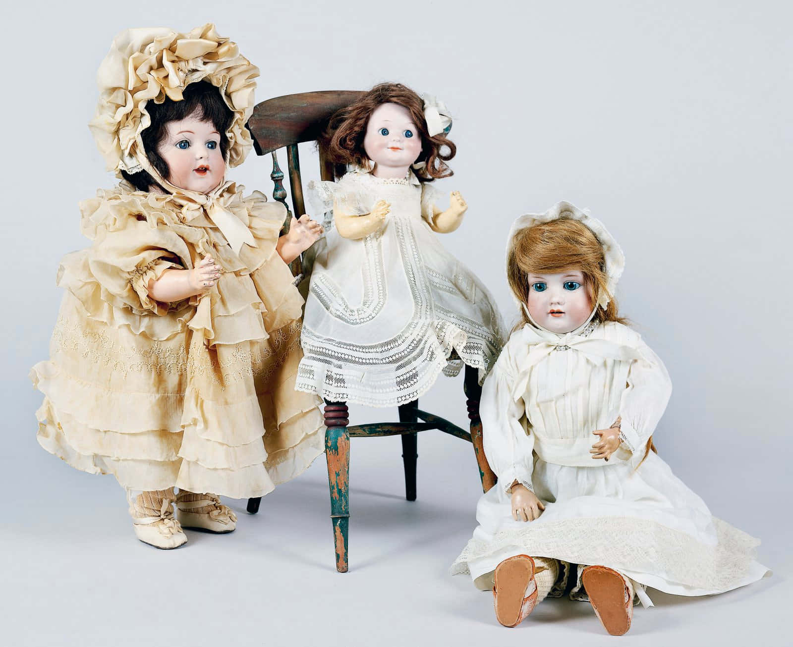 Dolls 桌布 壁紙 — 三個復古娃娃穿著精緻的奶油色和白色蕾絲連衣裙，在純白色背景前圍繞一張風化的木椅擺姿勢。