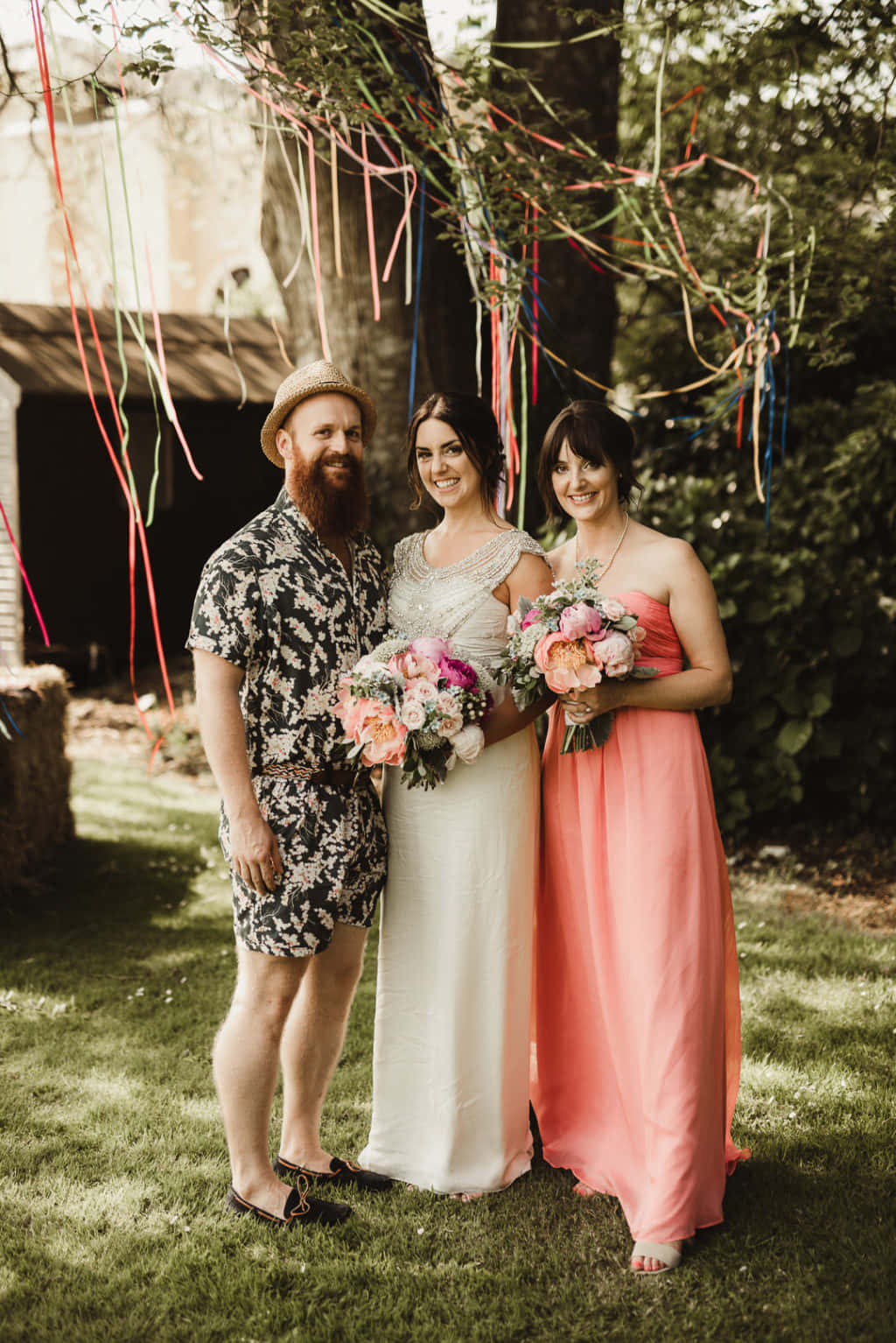 Bridal 桌布 壁紙 — 三個微笑的人穿著婚禮服飾，站在陽光普照的花園中色彩繽紛的絲帶下。