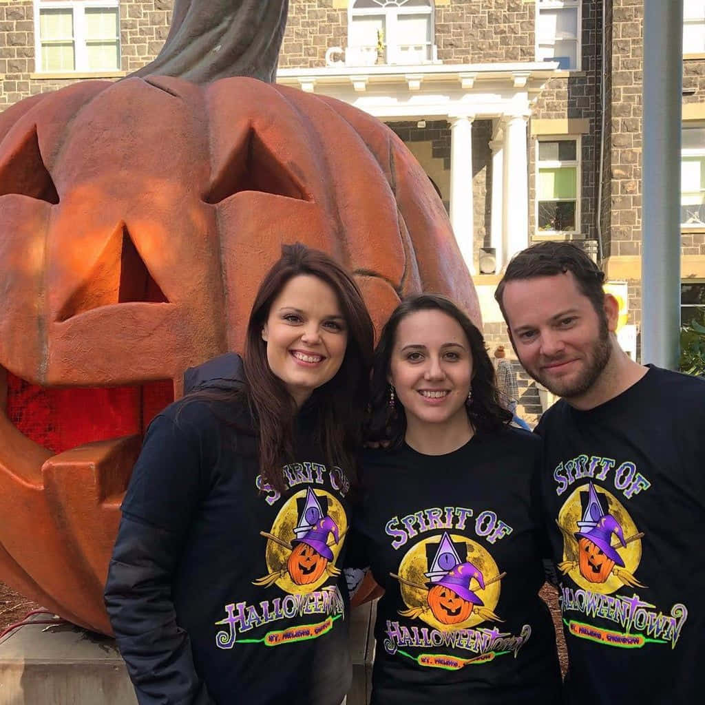 Halloweentown 桌布 壁紙 — 三個微笑的人穿著相配的萬聖節小鎮T恤，在一個巨大的發光傑克南瓜燈雕像前擺姿勢。