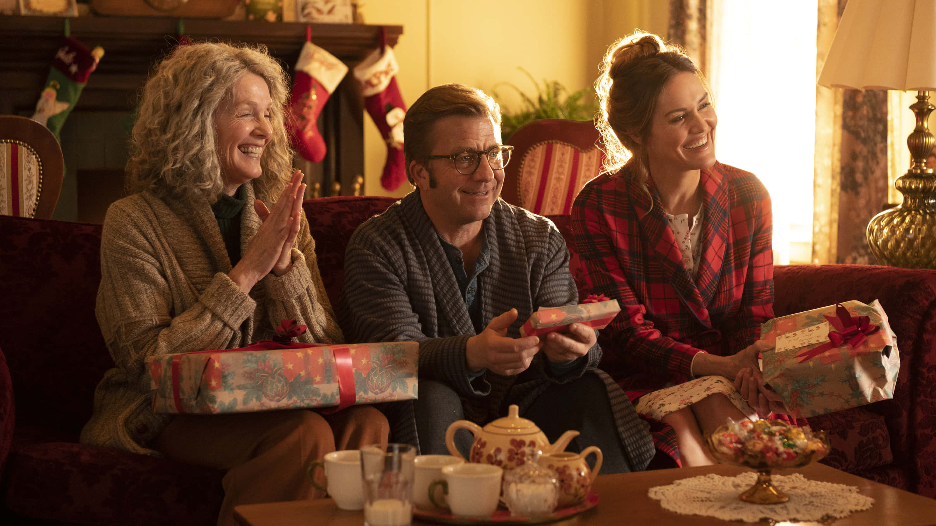a christmas story 桌布 壁紙 — 三個微笑的人穿著舒適的節日毛衣坐在紅色沙發上，拿著包裝好的禮物一起大笑。