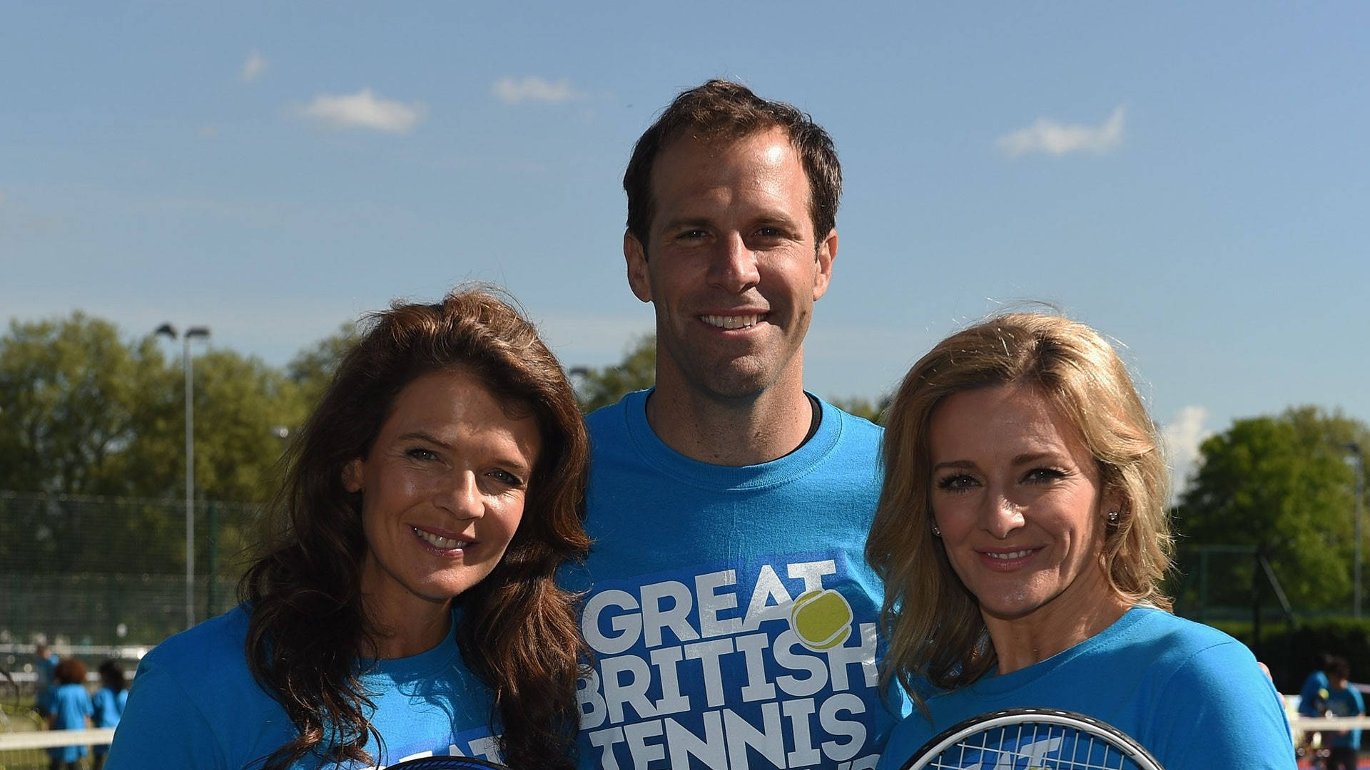 運動 桌布 壁紙 — 三個微笑的人穿著藍色「Great British Tennis」襯衫，在陽光明媚的戶外網球場上一起擺姿勢。