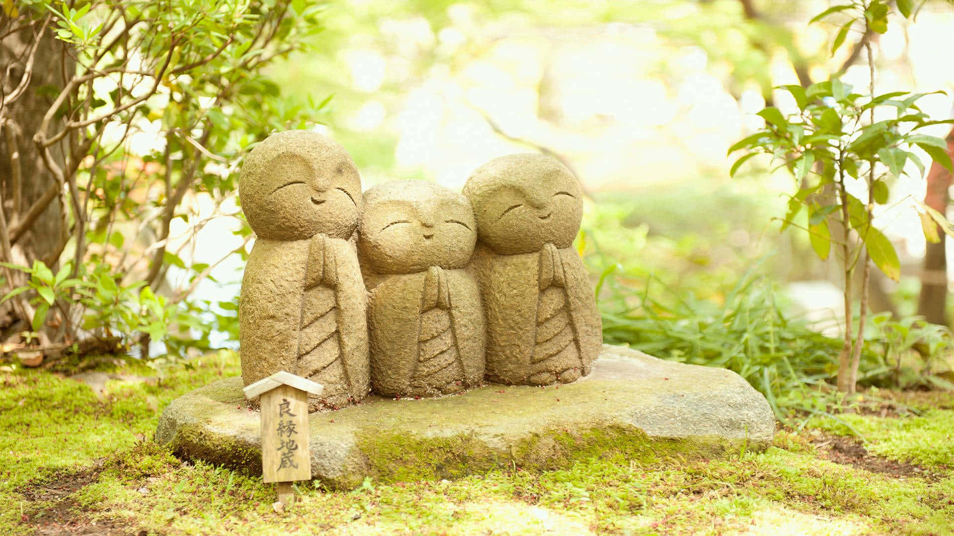 禪 桌布 壁紙 — 三個微笑的孩子石像一起坐在陽光普照的花園中一塊長滿苔蘚的石頭上，周圍綠意盎然。
