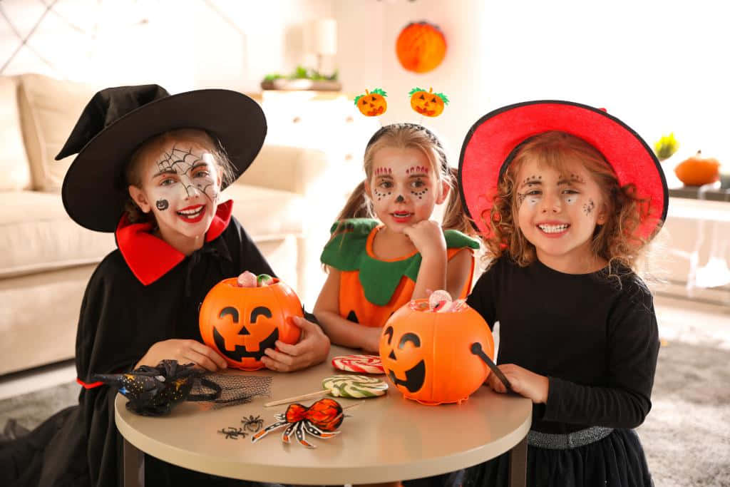 Halloween Pumpkin 桌布 壁紙 — 三個微笑的孩子穿著萬聖節服裝，在室內的桌子上拿著南瓜燈籠桶擺姿勢。