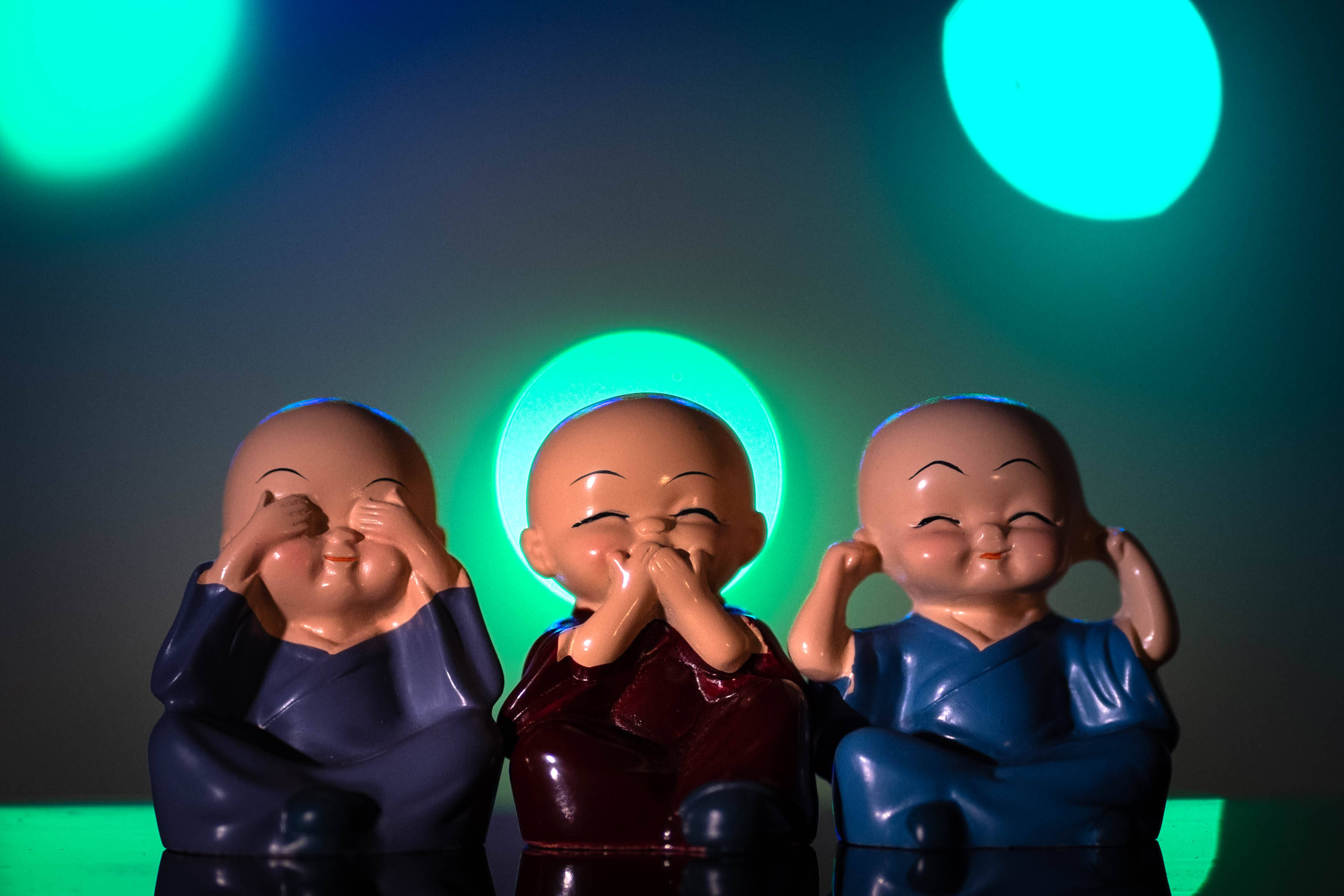 Dolls 桌布 壁紙 — 三個微笑的陶瓷僧侶雕像遮住眼睛、耳朵和嘴巴，背景是發光的青綠色散景燈光。