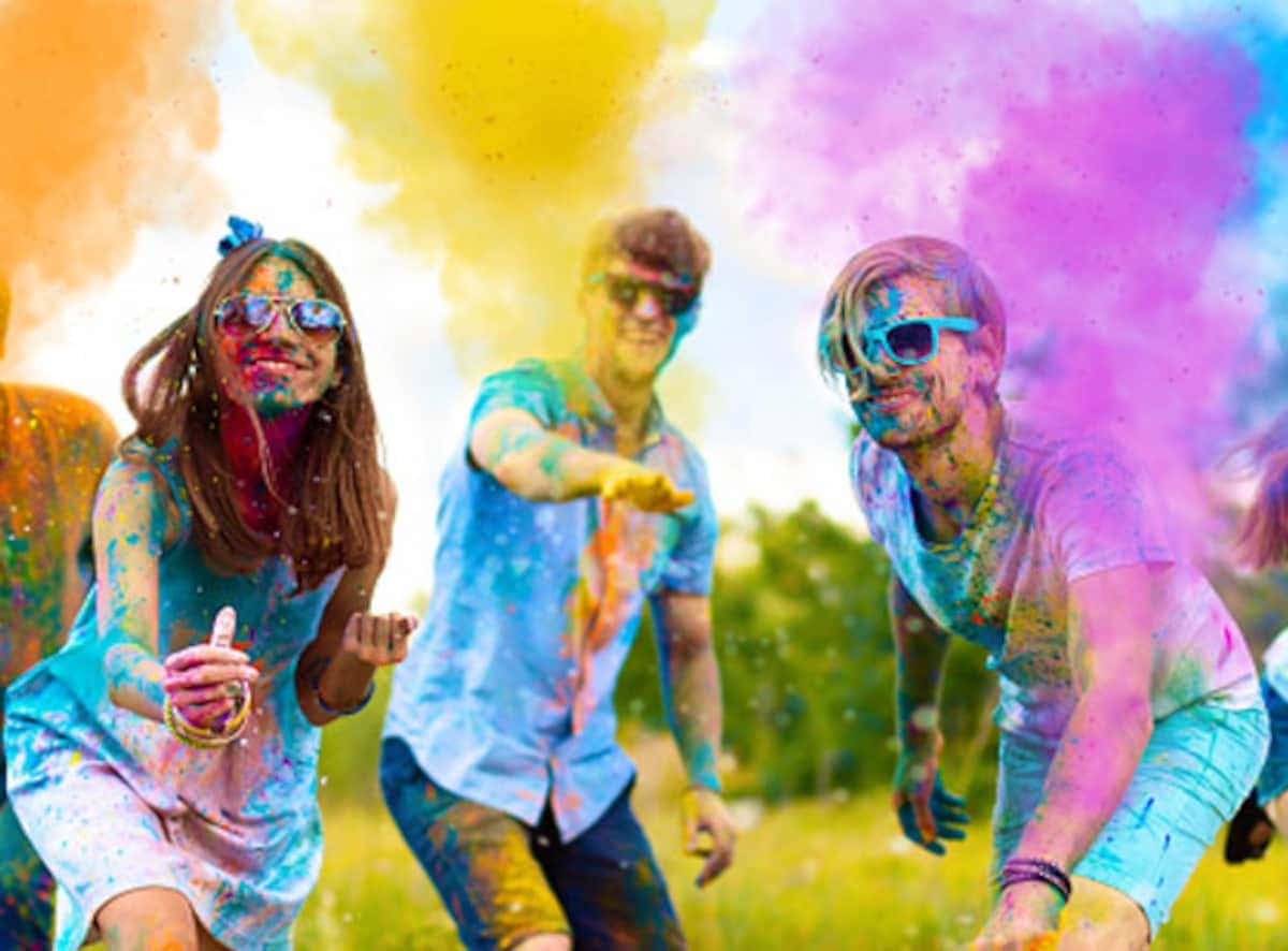 Holi 桌布 壁紙 — 三個快樂的人身上沾滿鮮豔的粉末，在陽光明媚的田野中跳躍大笑，慶祝侯麗節。