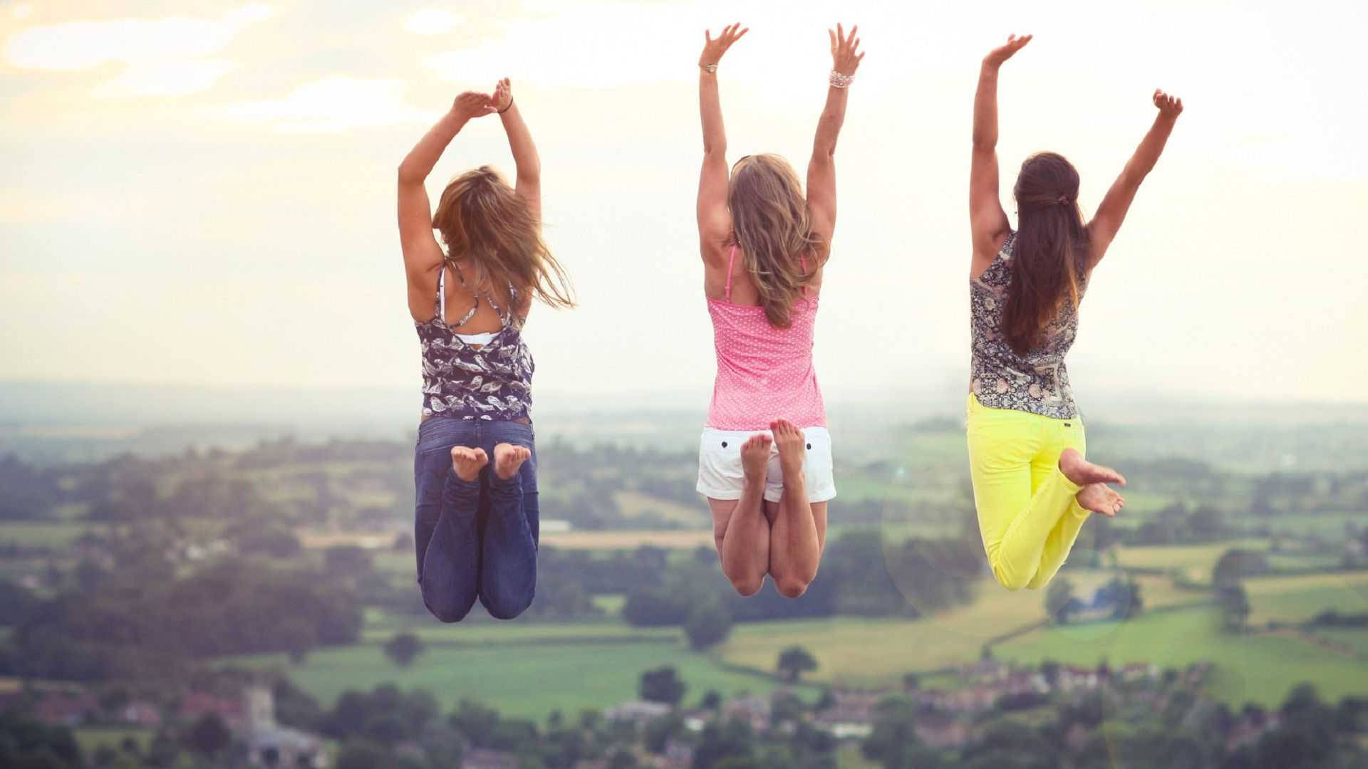 3 Best Friends 桌布 壁紙 — 三個快樂的女孩在廣闊的綠色山谷上空，舉起雙臂，在柔和的金色夕陽映襯下騰空跳躍。