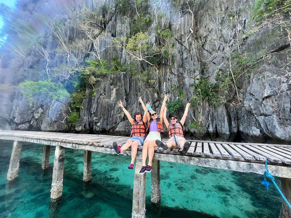 Travel Desktop 桌布 壁紙 — 三個快樂的朋友穿著救生衣坐在木碼頭上，高舉雙臂，背景是高聳的石灰岩懸崖和碧綠的海水。