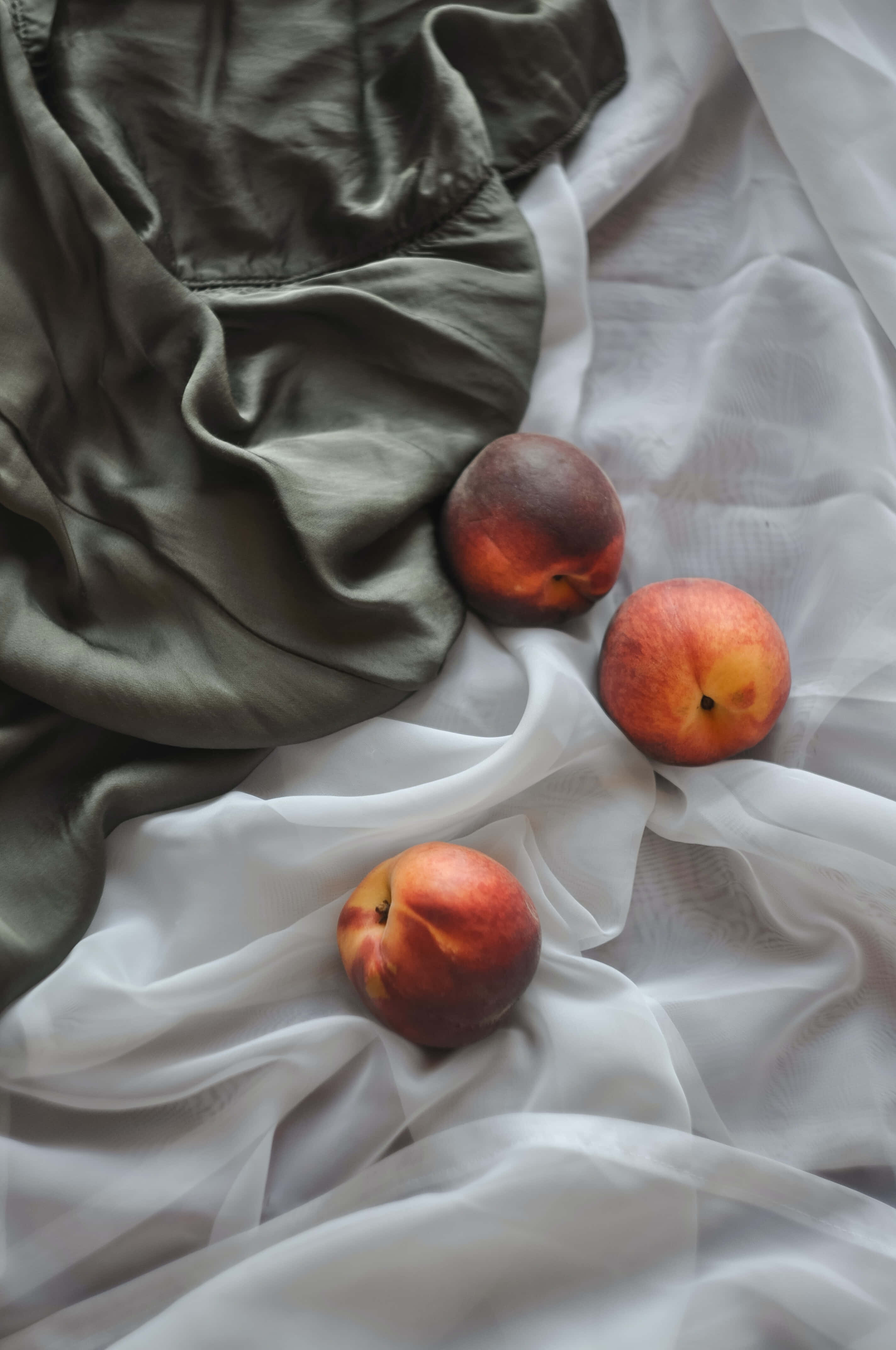 Peaches 桌布 壁紙 — 三個成熟的桃子放在皺起的白色和橄欖綠色布料上，在柔和自然光下。