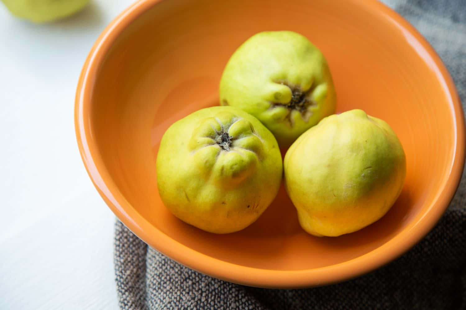 Quince 桌布 壁紙 — 三個成熟的榲桲放在一個溫暖的橙色陶瓷碗中，置於有紋理的灰色布上。