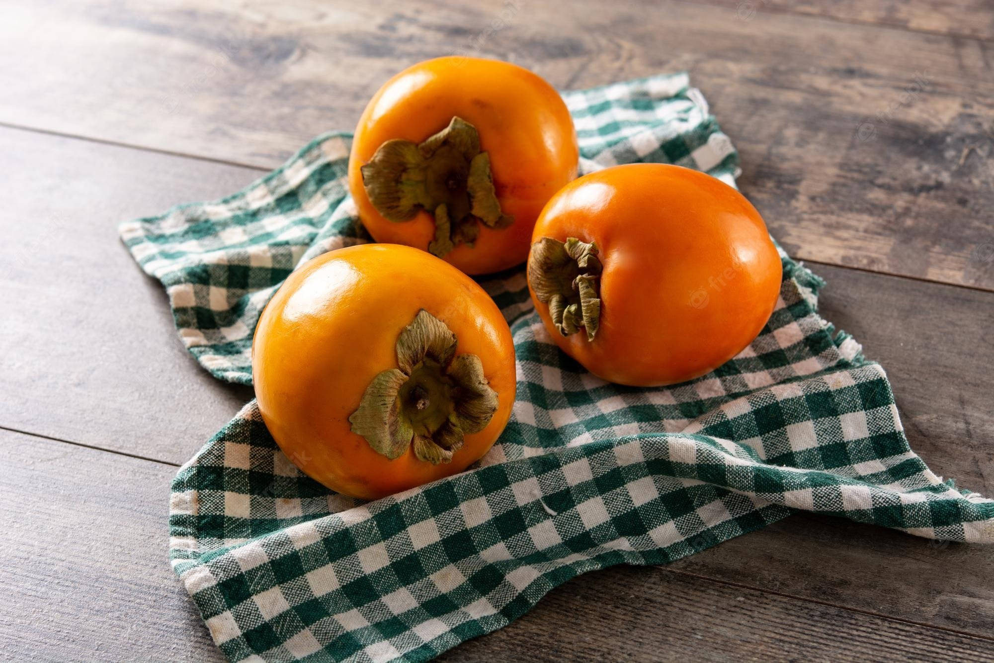Fruit 桌布 壁紙 — 三個成熟的橙色柿子放在一塊綠色和白色格子布上，置於經風化的木質表面。