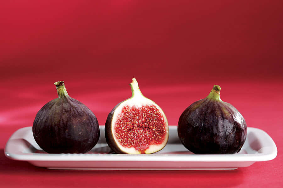 Purple Figs 桌布 壁紙 — 三個成熟的無花果，其中一個切開露出多汁的紅色果肉，放在白色盤子上，背景為鮮紅色。