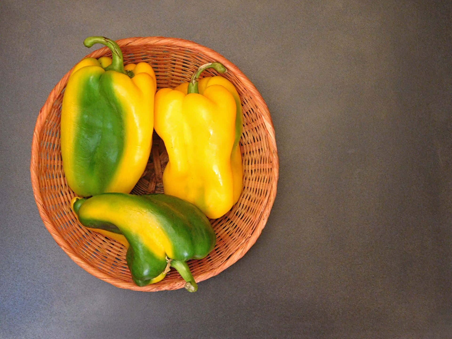 Peppers 壁紙 — 三個成熟的黃色和綠色甜椒放在一個編織的柳條籃子裡，置於深灰色表面上。