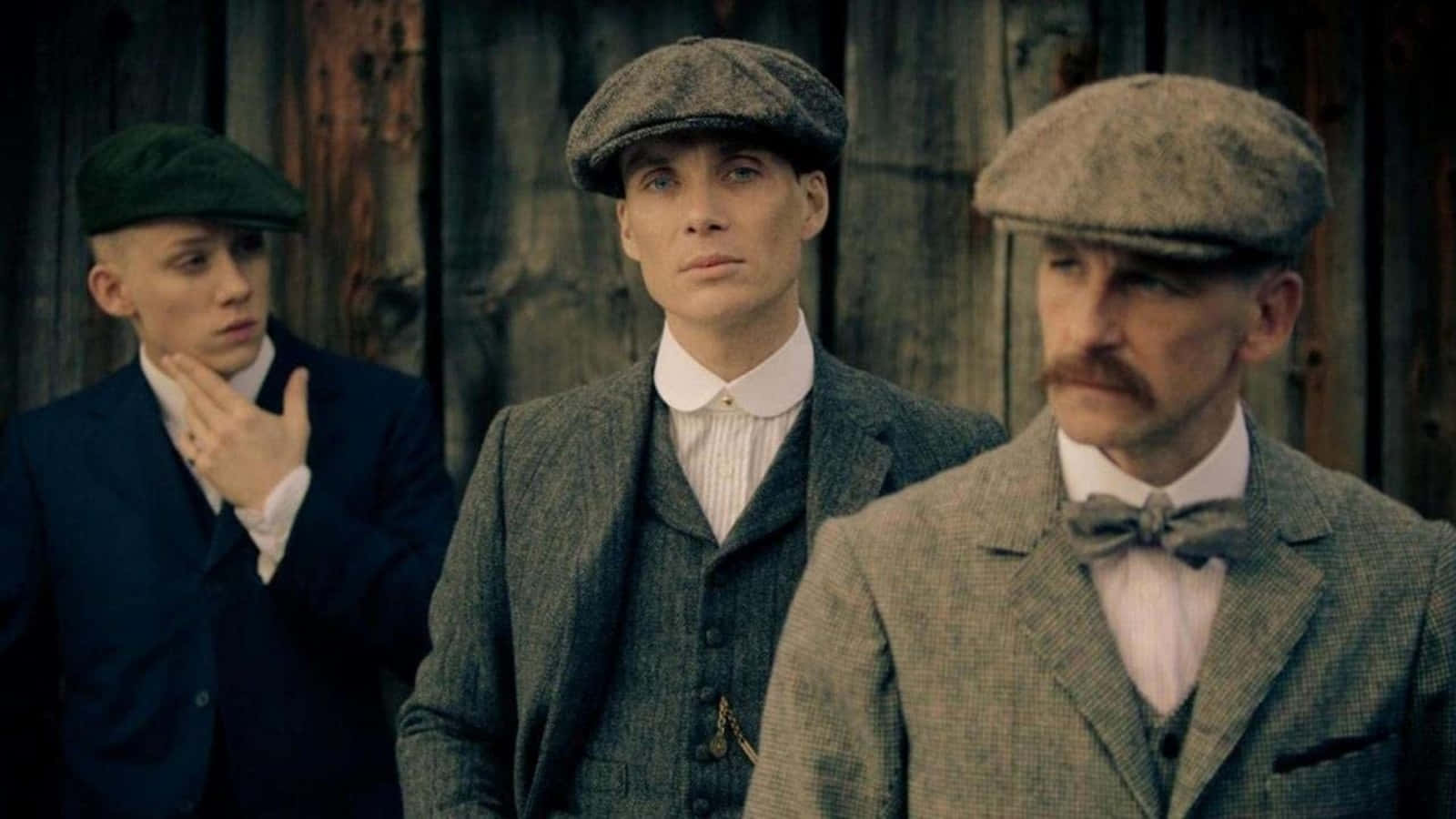 三個身穿復古西裝、戴著平頂帽的男人，莊嚴地站在一面木牆前。 — Peaky Blinders 壁紙
