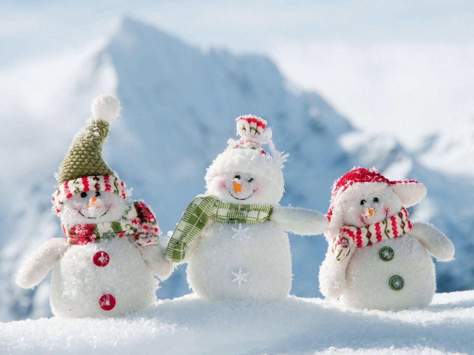 Cute Snow 桌布 壁紙 — 三個戴著節日帽子和圍巾的快樂雪人公仔在明亮的陽光下一起站在白雪覆蓋的山坡上。