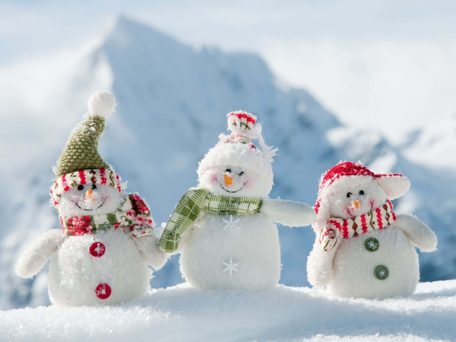 可愛聖誕節 桌布 壁紙 — 三個戴著節日帽子和圍巾的快樂雪人，在明亮的陽光下一起站在白雪覆蓋的山坡上。