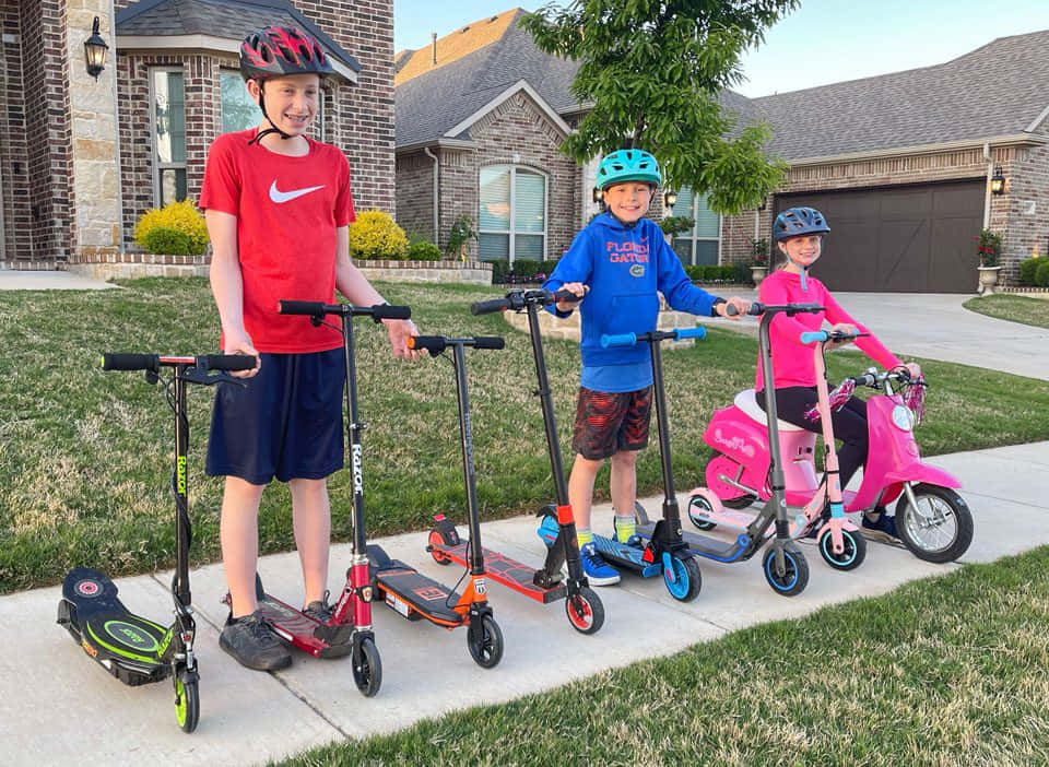 scooter 桌布 壁紙 — 三個戴頭盔的笑臉小孩在郊區人行道上，於黃金時刻與彩色滑板車一起擺姿勢。