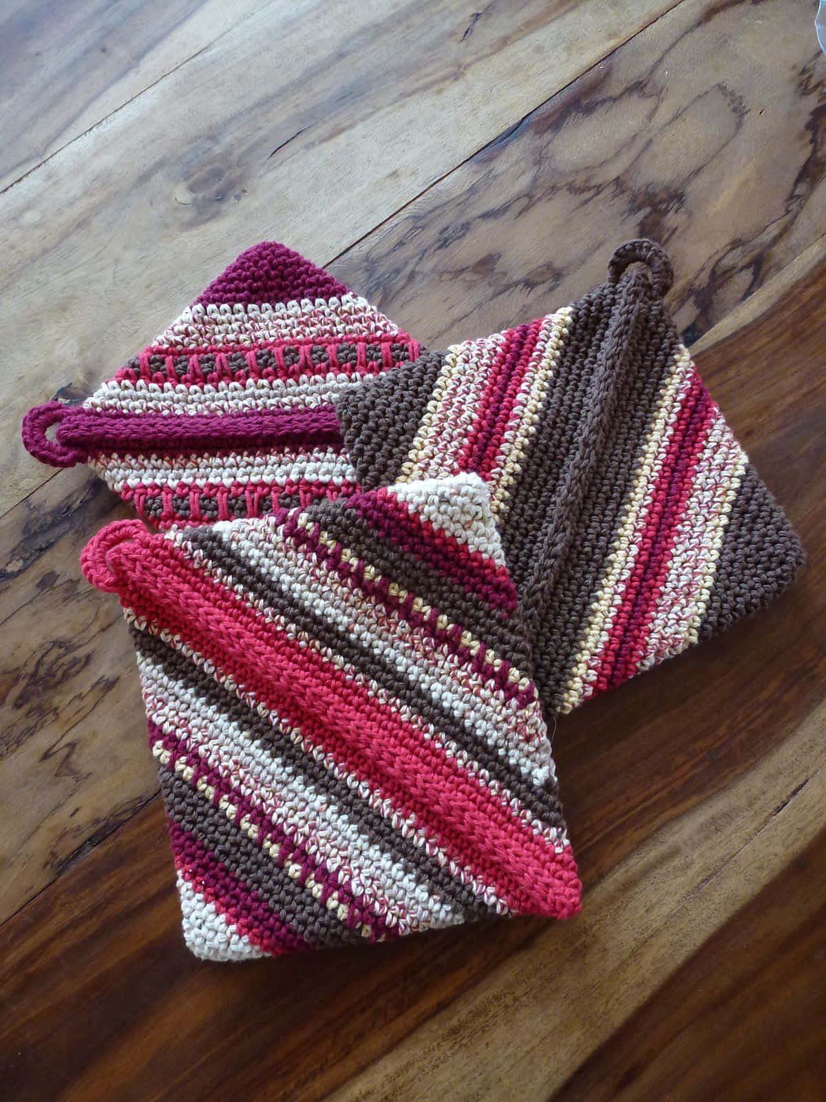 Crochet 桌布 壁紙 — 三個手織隔熱墊，帶有粉紅、棕色和奶油色的斜紋，放在一張質樸的木桌上。