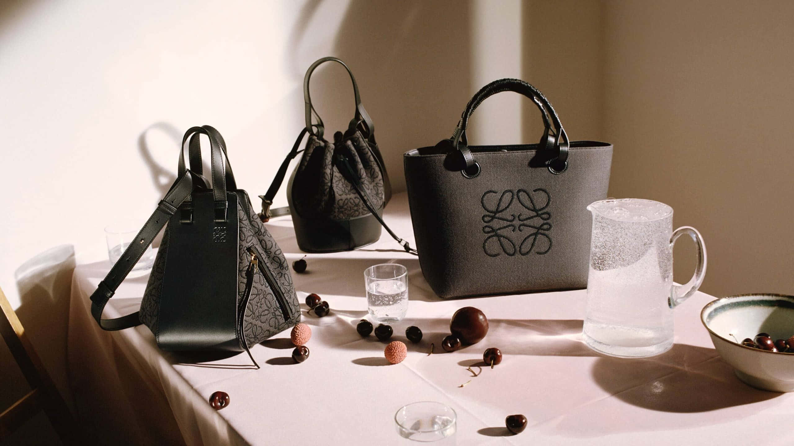 Luxury Brands 桌布 壁紙 — 三個時尚的灰色Loewe手提包擺放在陽光照射的桌子上，散落著櫻桃和玻璃器皿。