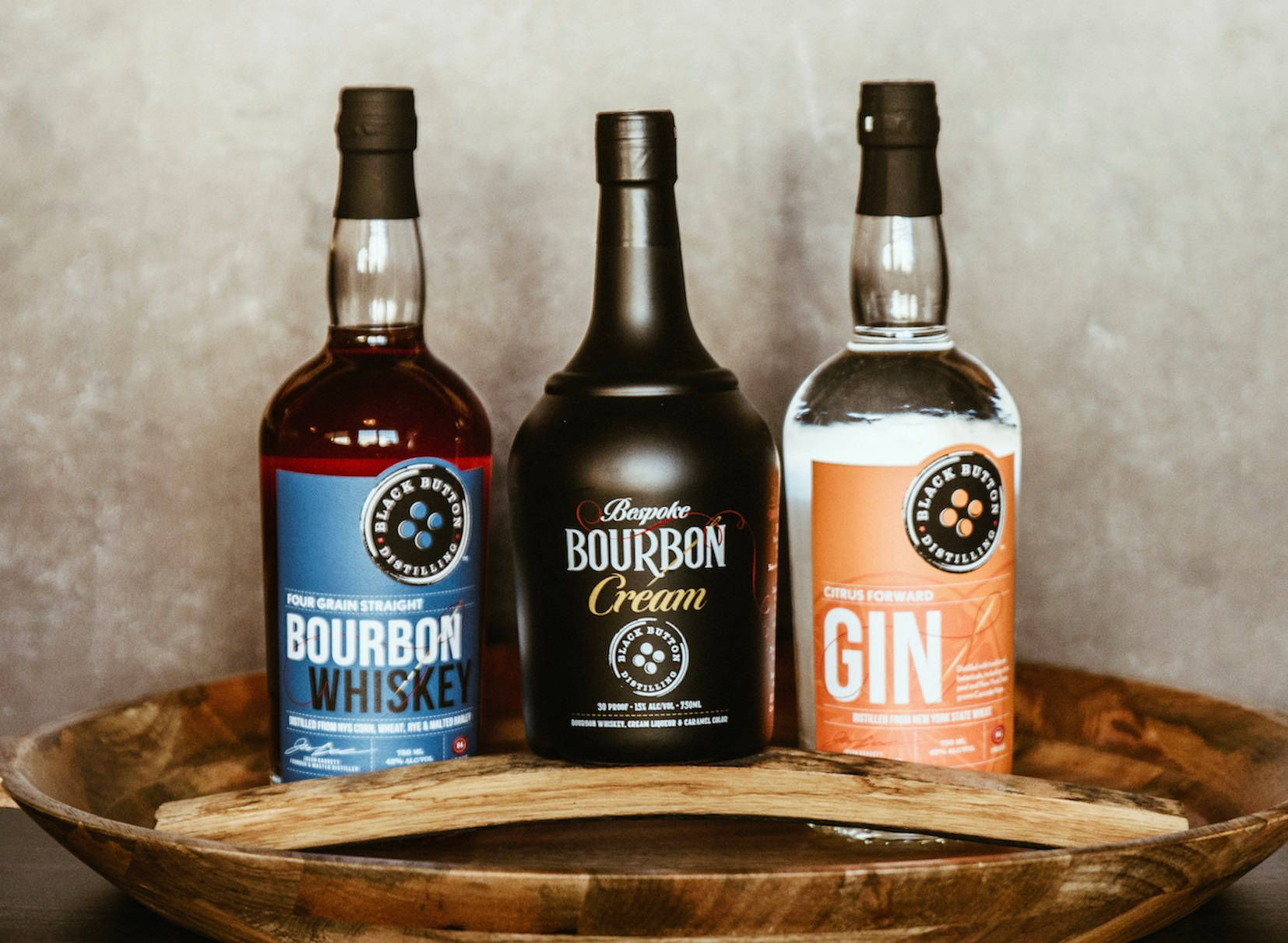alcohol 桌布 壁紙 — 三個有標籤的酒瓶（波本威士忌、波本奶油、琴酒）優雅地放置在一個質樸的木碗中，背景是帶紋理的米色牆壁。