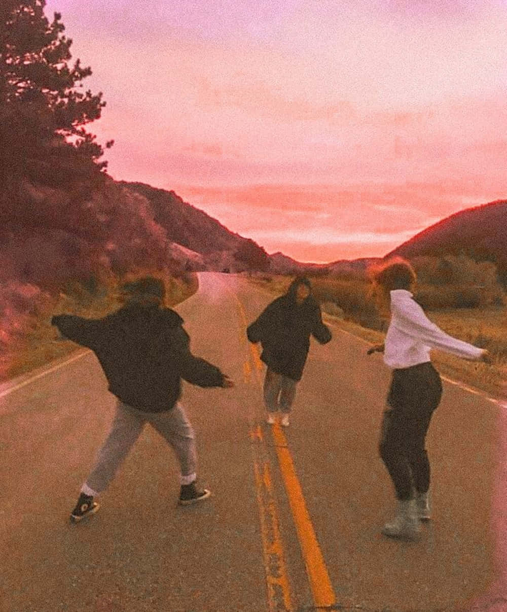 Best Friend Aesthetic 桌布 壁紙 — 三個朋友在夢幻粉紅色的日落天空下，沿著蜿蜒的山路歡快地奔跑。