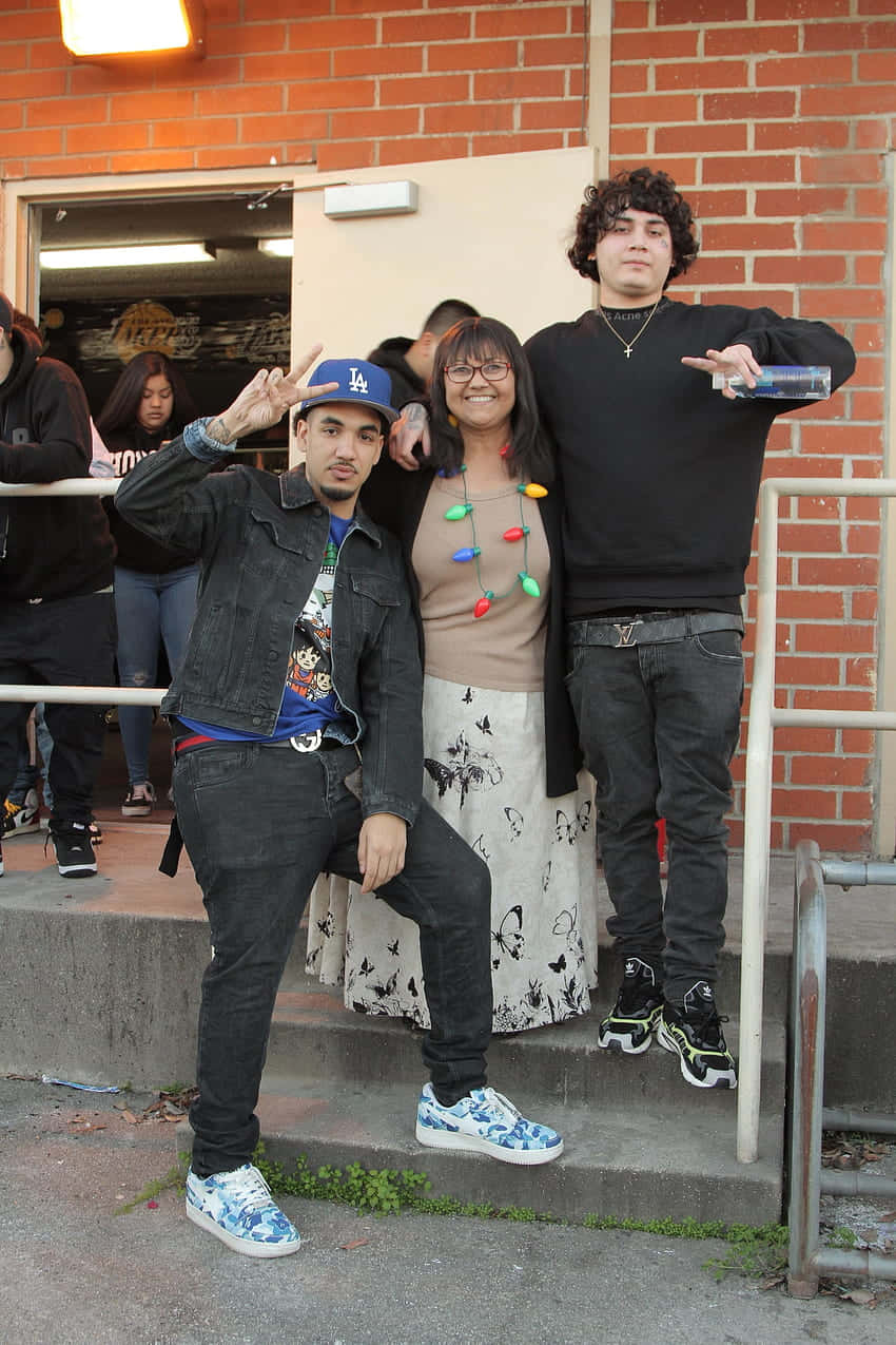 Shoreline Mafia 桌布 壁紙 — 三個朋友在磚砌建築外的混凝土階梯上愉快地擺姿勢。其中一人戴著捲曲假髮並拿著水瓶。