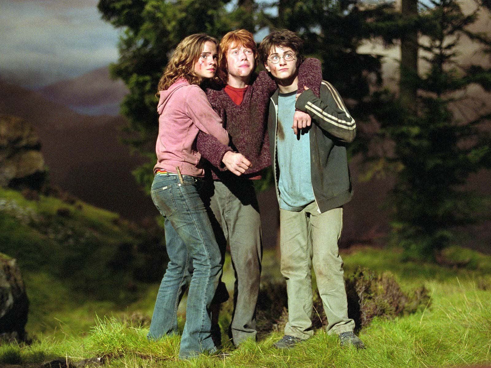 Golden Trio 桌布 壁紙 — 三個朋友在長滿草的山坡上緊密地站在一起，背景有樹木和遠處的山脈，天空多雲。