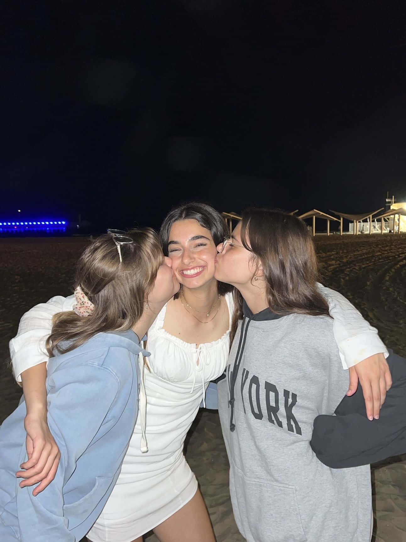 Friends 桌布 壁紙 — 三個朋友在黑暗的海灘上大笑並親吻臉頰，遠處有發光的藍色燈光。