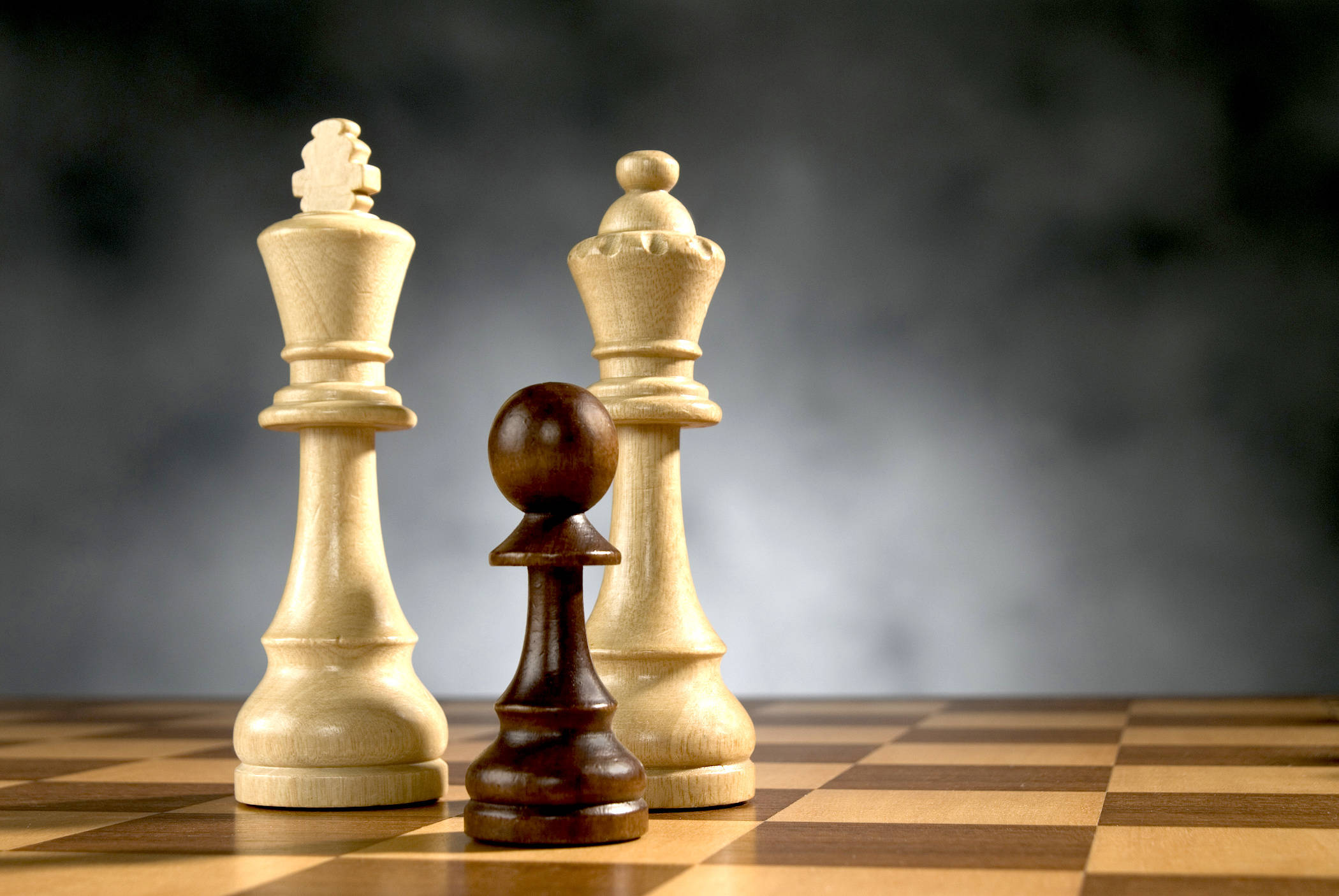 運動 桌布 壁紙 — 三個木製國際象棋棋子，一個深色兵卒兩側有兩個淺色國王，站在棋盤上，背景為煙灰色。