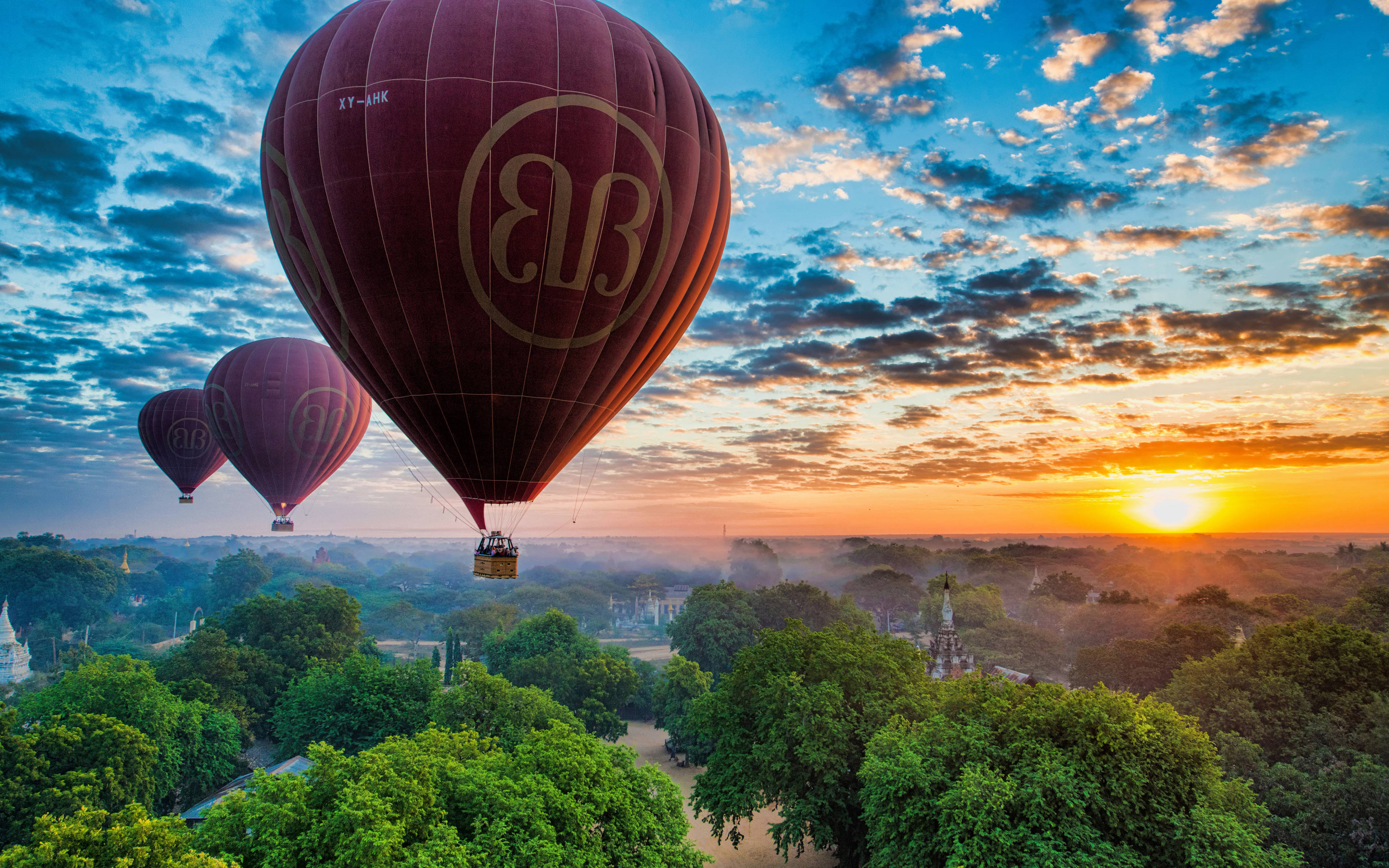 Hot Air Balloon 桌布 壁紙 — 三個栗色熱氣球在日出時分飄浮在茂密的綠色森林上空，金色的光芒塗抹著天空。