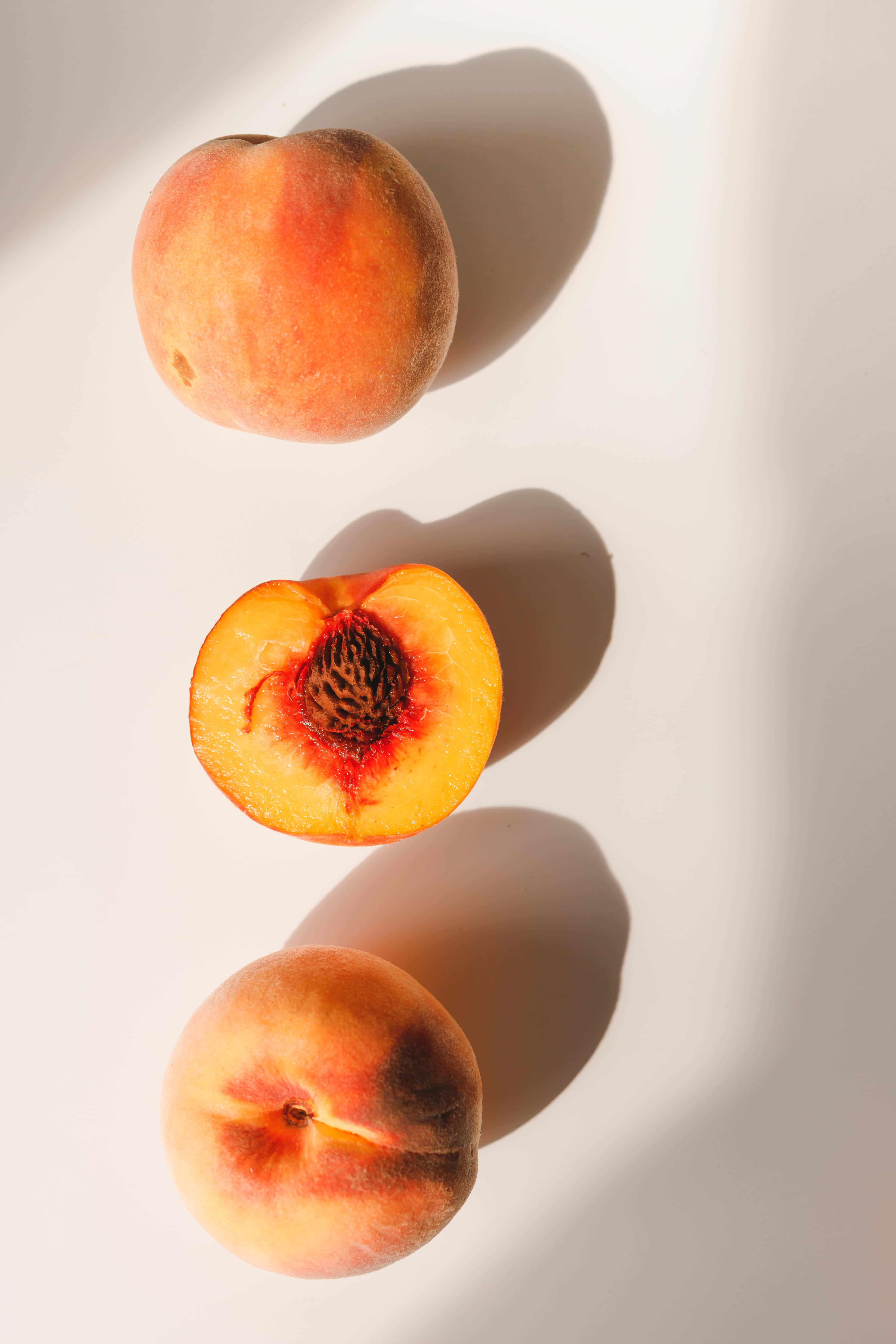 Peaches 桌布 壁紙 — 三個桃子，其中一個切半，在溫暖陽光下於明亮的白色表面上投下柔和陰影。