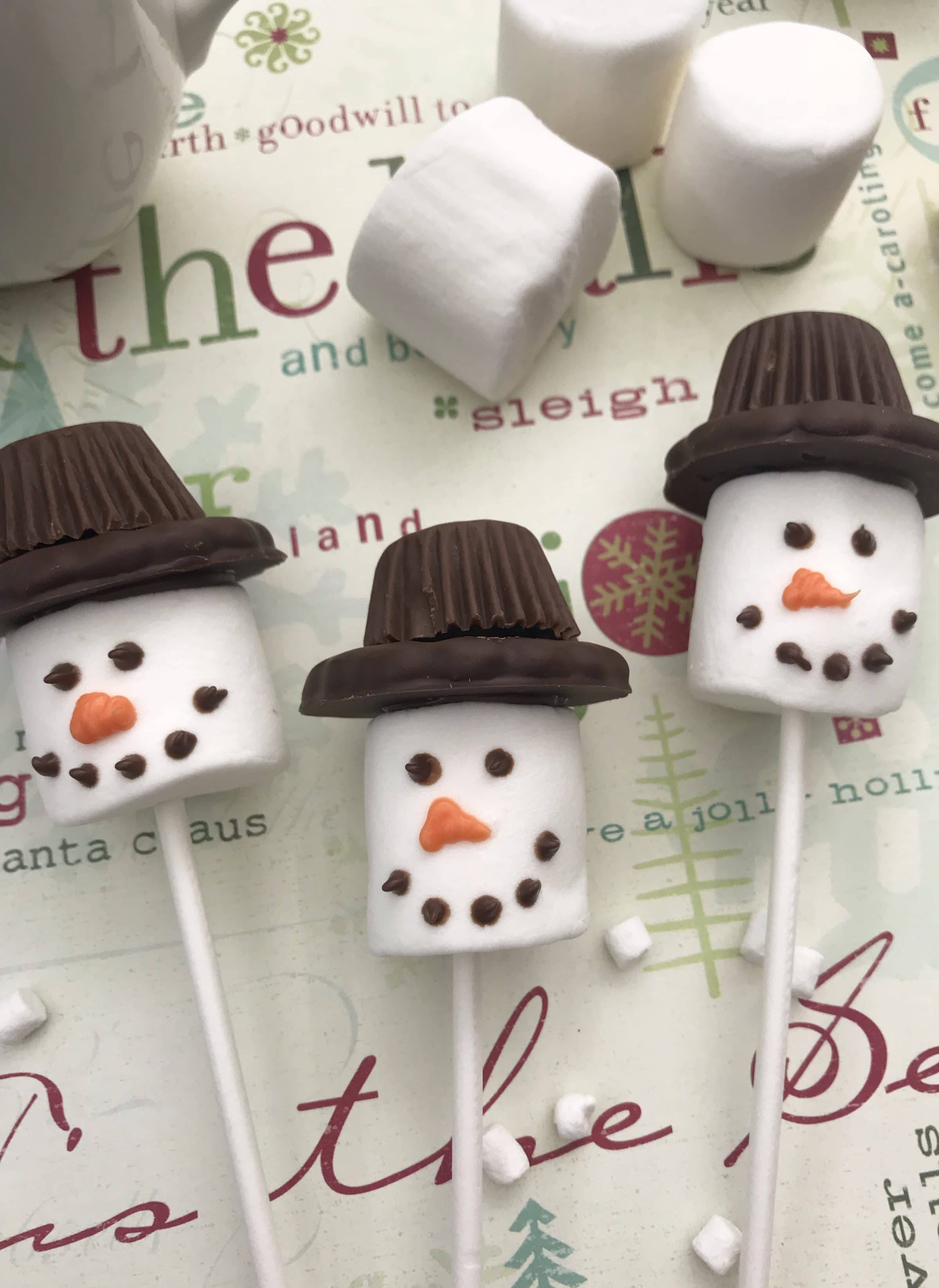 Marshmallow 桌布 壁紙 — 三個棉花糖雪人插在棍子上，戴著巧克力帽子和糖果臉，放在節日主題的紙背景上。