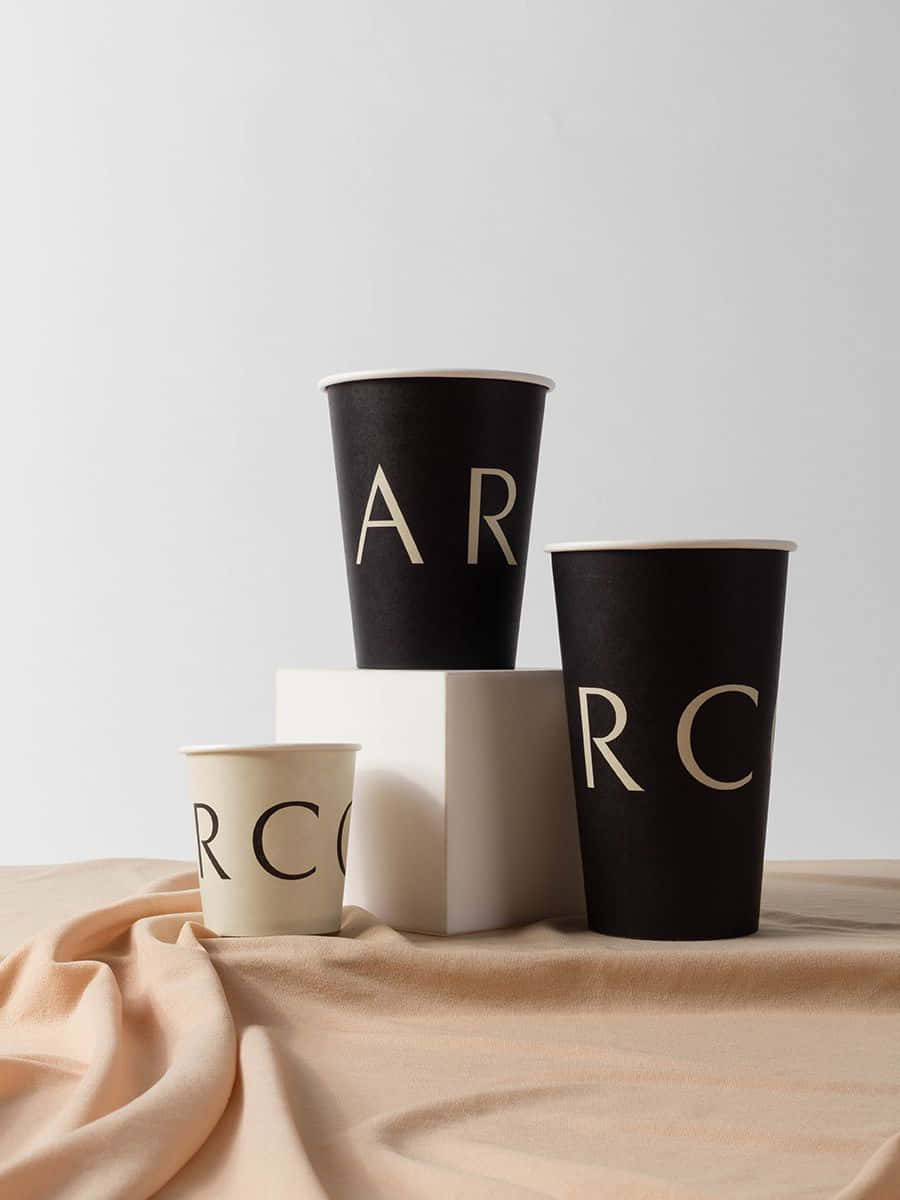 Minimalist Coffee 桌布 壁紙 — 三個極簡風格品牌紙杯，帶有金色字樣，排列在米色布料上，背景為純白色。