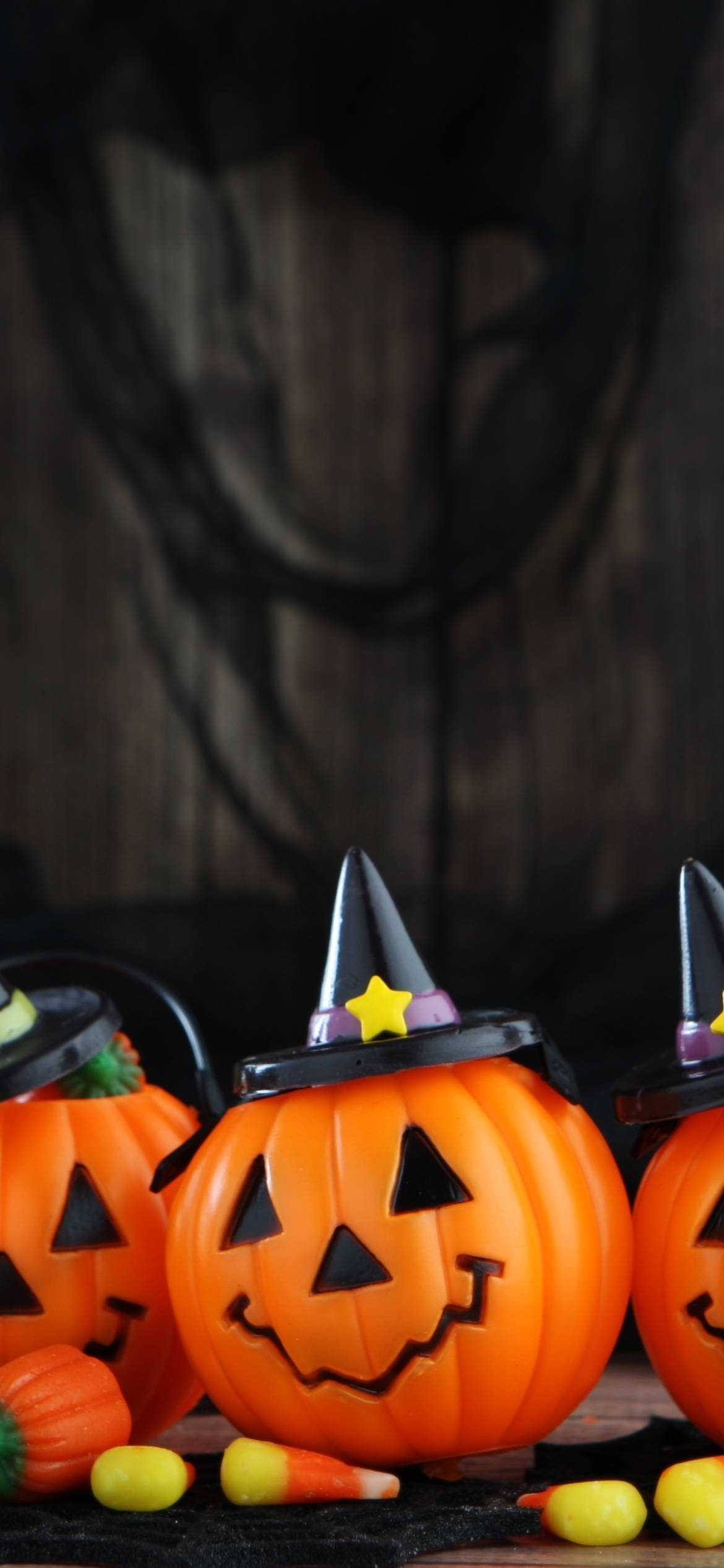 Cute Halloween Phone 桌布 壁紙 — 三個橙色塑膠南瓜，戴著巫婆帽和傑克南瓜燈臉孔，放在黑色布上，旁邊有糖果玉米和深色木質背景。