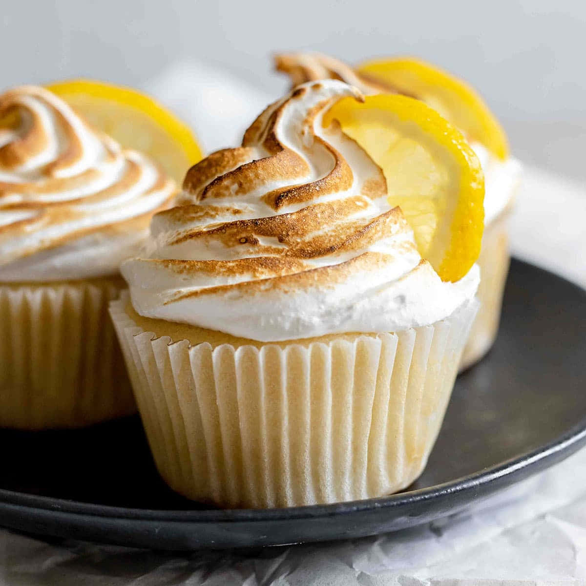 Meringue 桌布 壁紙 — 三個檸檬蛋白霜杯子蛋糕，帶有烤焦的頂峰和亮黃色切片，放在深色盤子上，背景是柔和的白色。