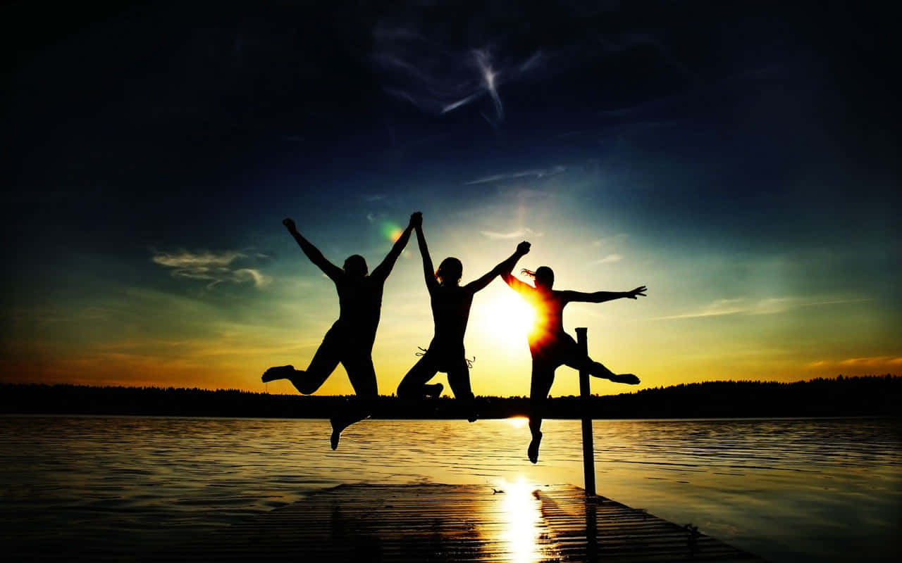 Friendship 桌布 壁紙 — 三個歡樂的剪影在碼頭上一起跳躍，夕陽在他們身後落下，將金色光芒灑在水面上。